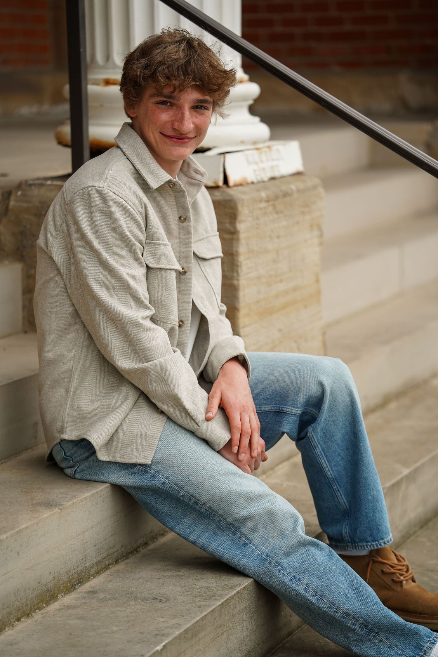 A person in a tan jacket sits casually on concrete steps wearing light wash jeans and brown boots.
