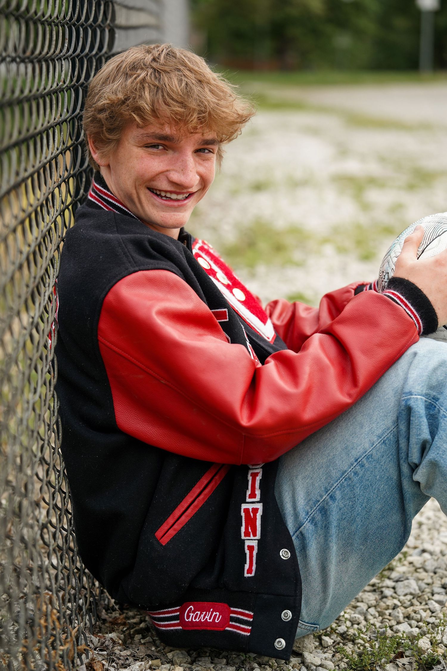 Student in red and black letterman jacket posing against chain link fence outdoors.