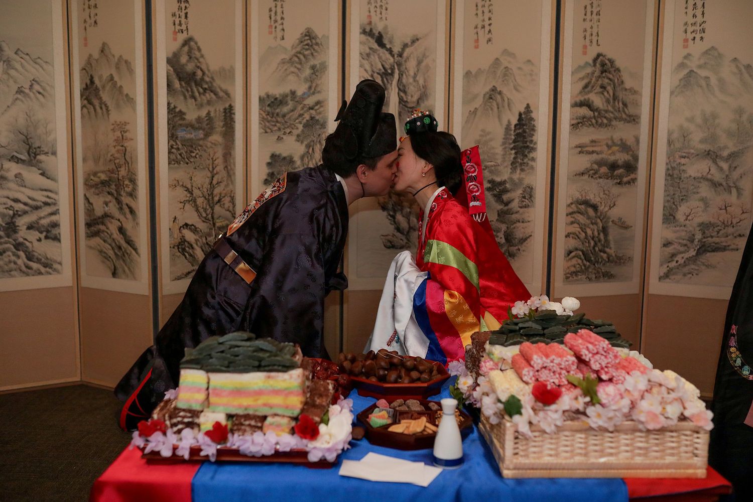 Traditional Korean ceremony participants arrange and fold colorful paper flowers at decorated tables with folding screens in background.