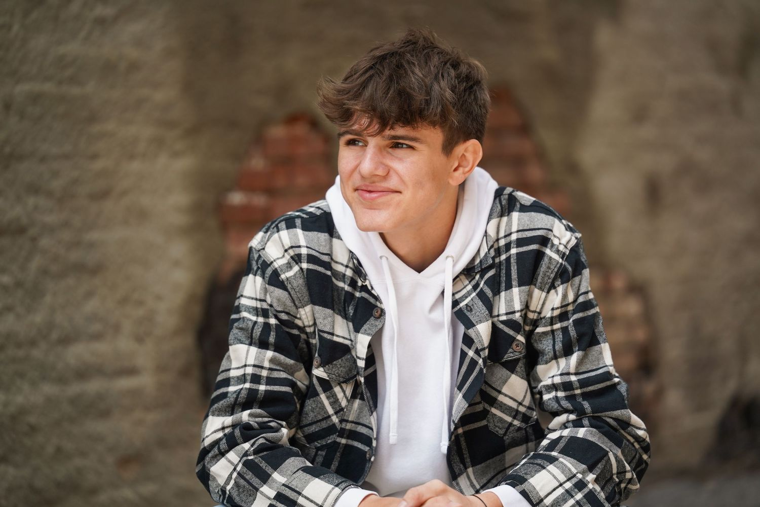 A person wearing a plaid flannel jacket and white t-shirt stands against a stone wall background.