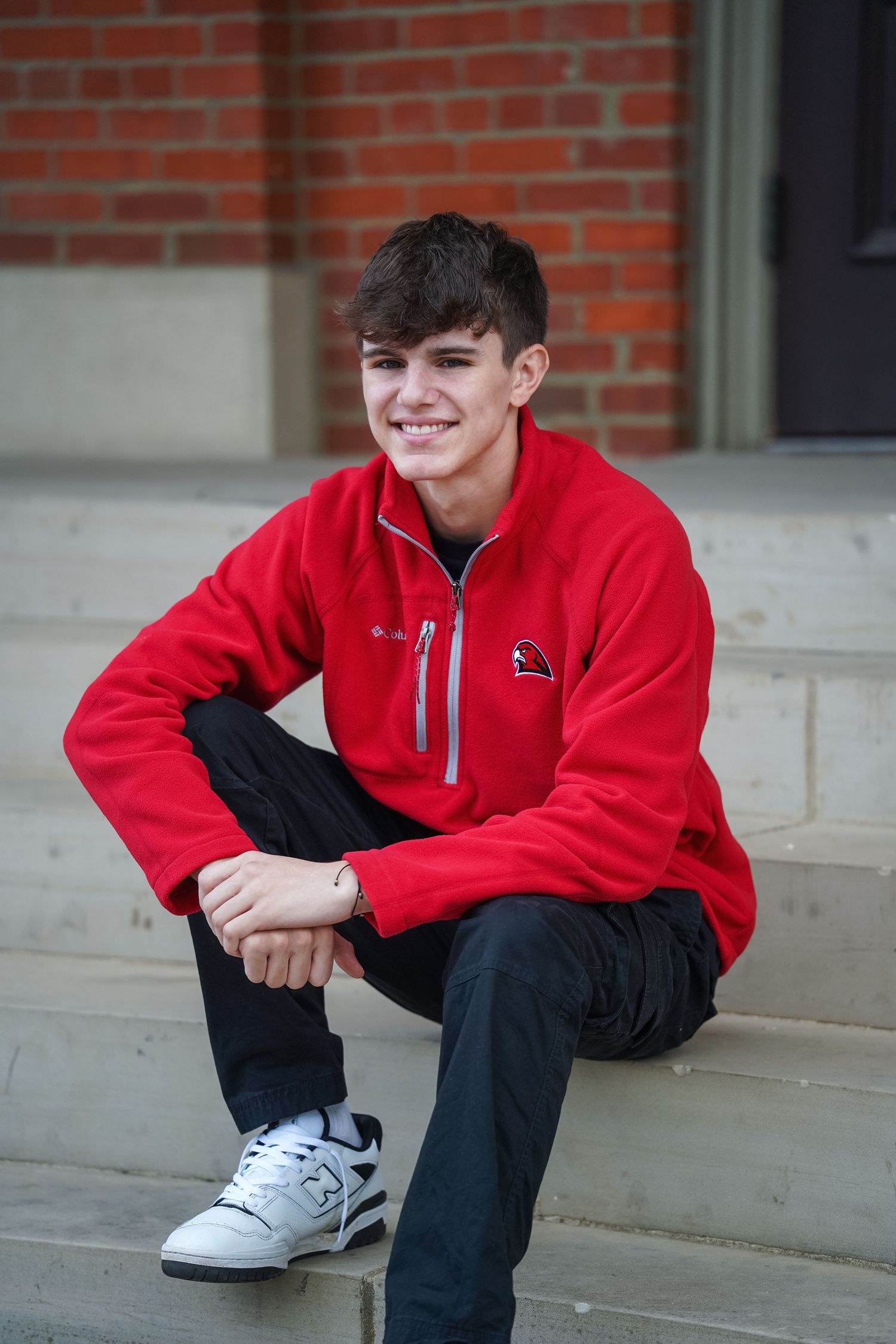 A person wearing a bright red fleece jacket and dark jeans sits casually on concrete steps near a brick wall.