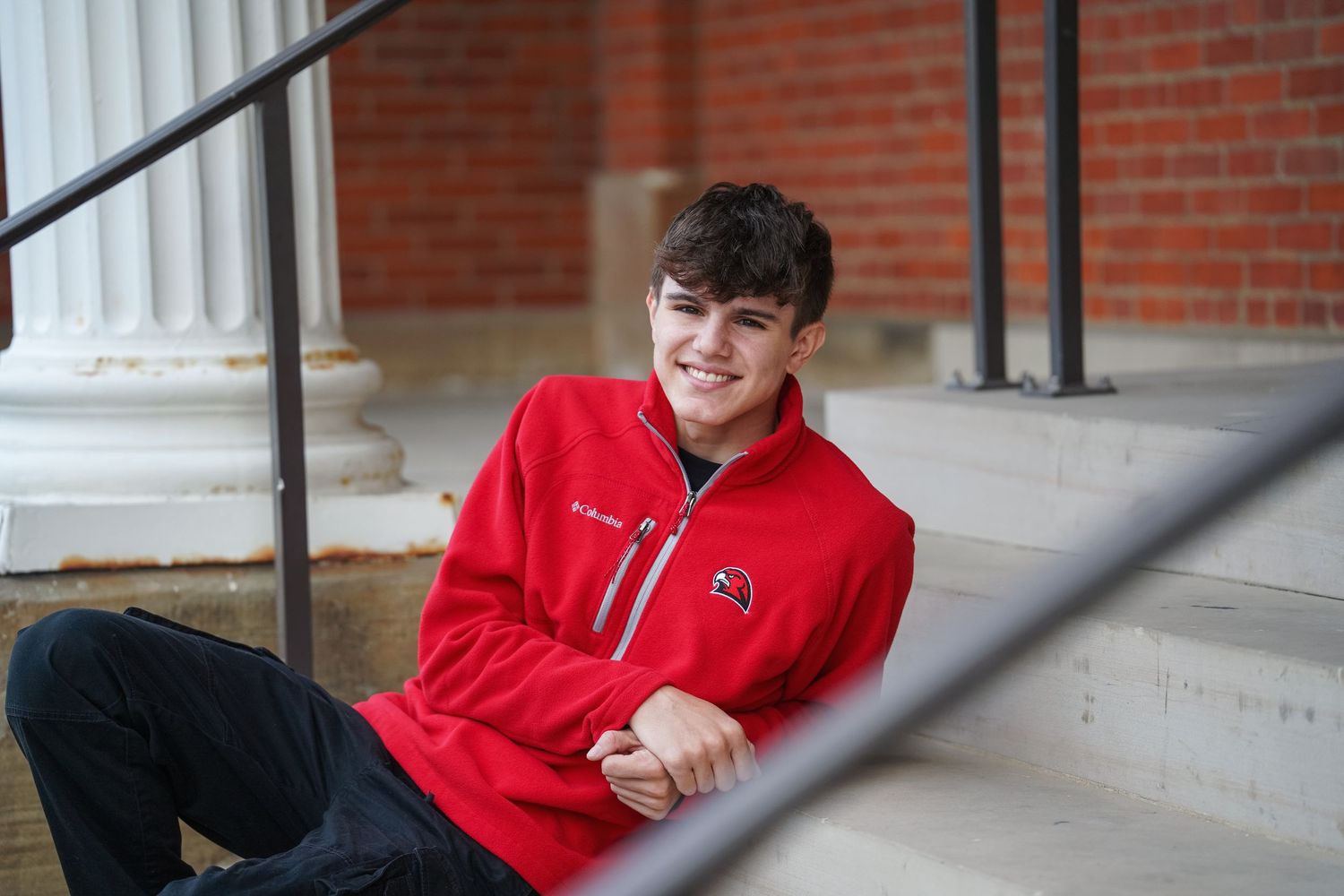 A student wearing a bright red hoodie sits casually on concrete steps with a brick wall backdrop.