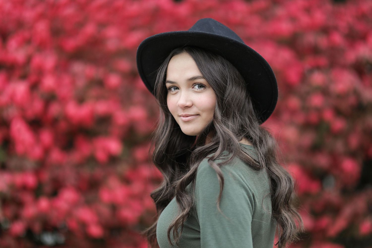 A person in a wide-brimmed black hat and sage green sweater poses against vibrant pink autumn foliage.