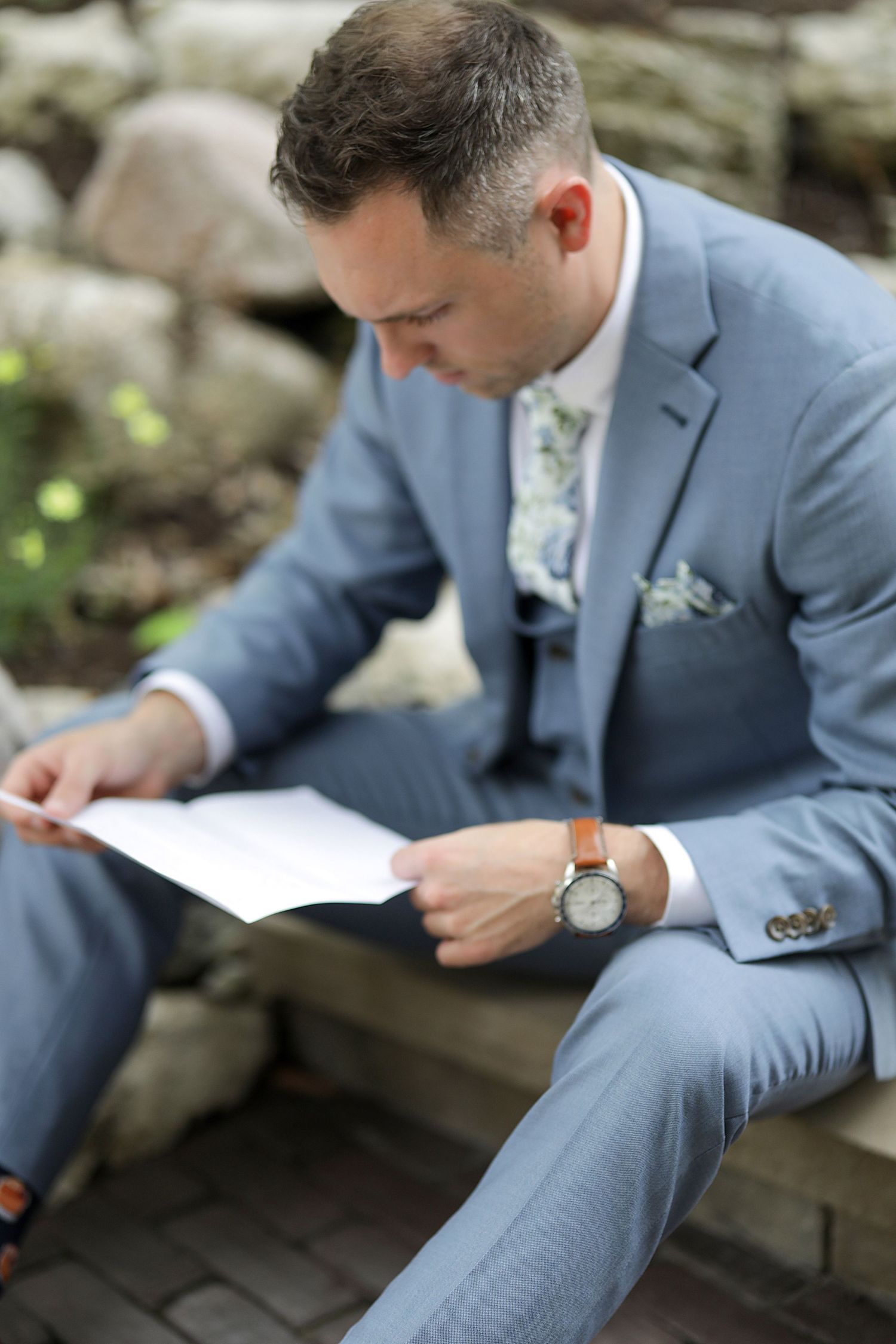 Groom in light blue suit reads letter while sitting outdoors wearing a floral tie and wristwatch.