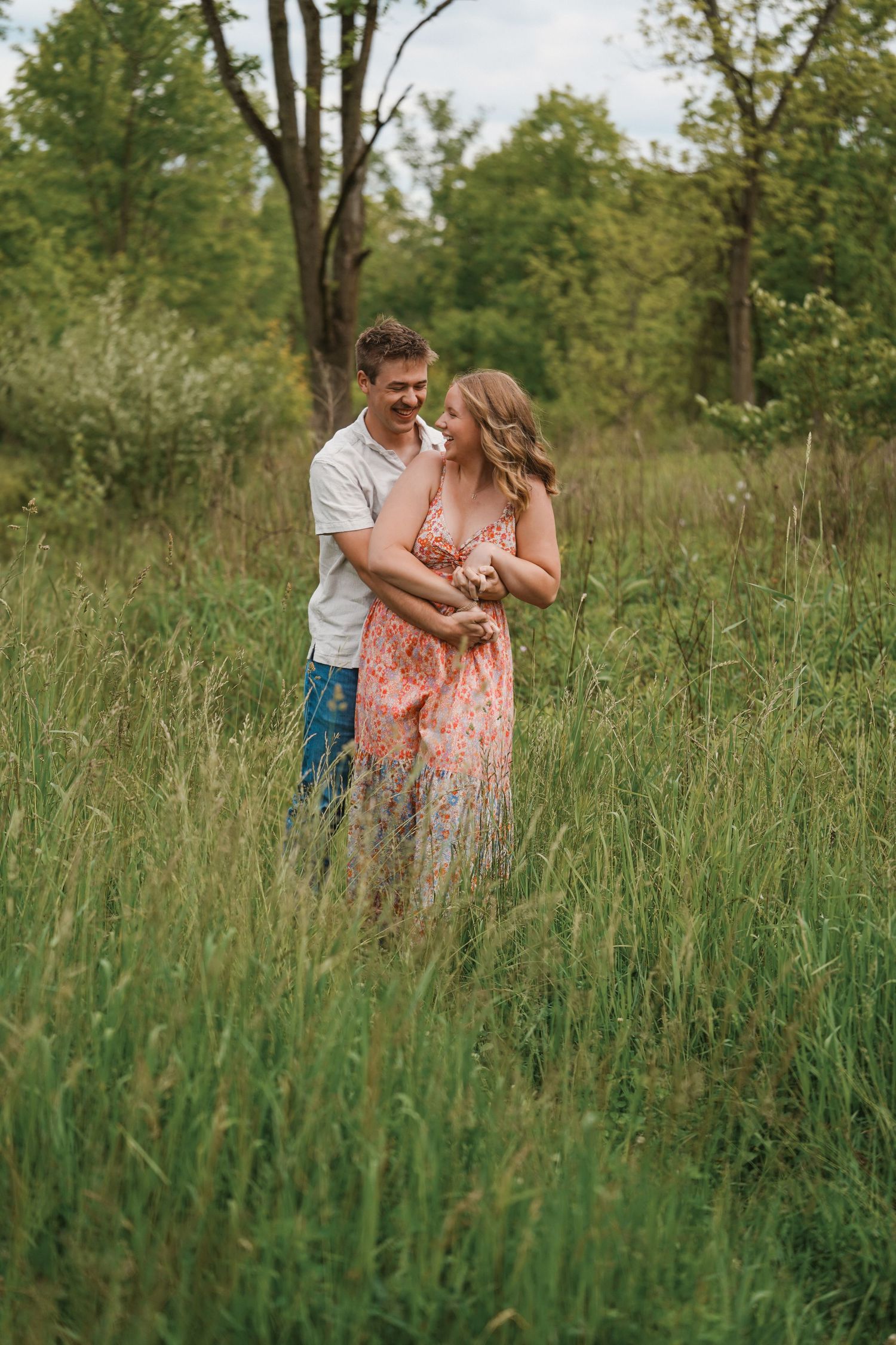 A couple shares a romantic moment in tall summer grass with sunlit trees in the background.