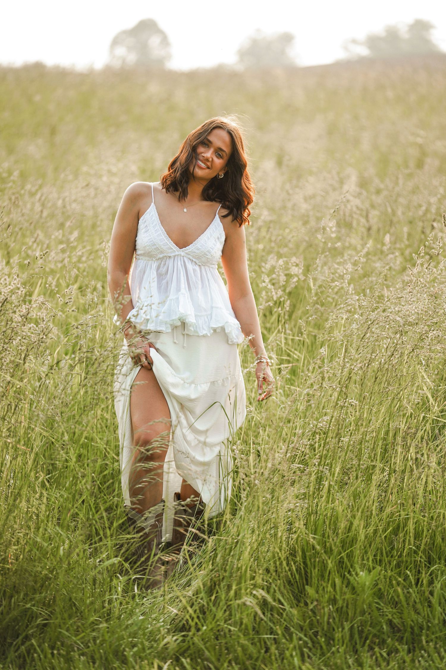 A person in a flowing white sundress walks through tall summer grass at golden hour in a field.