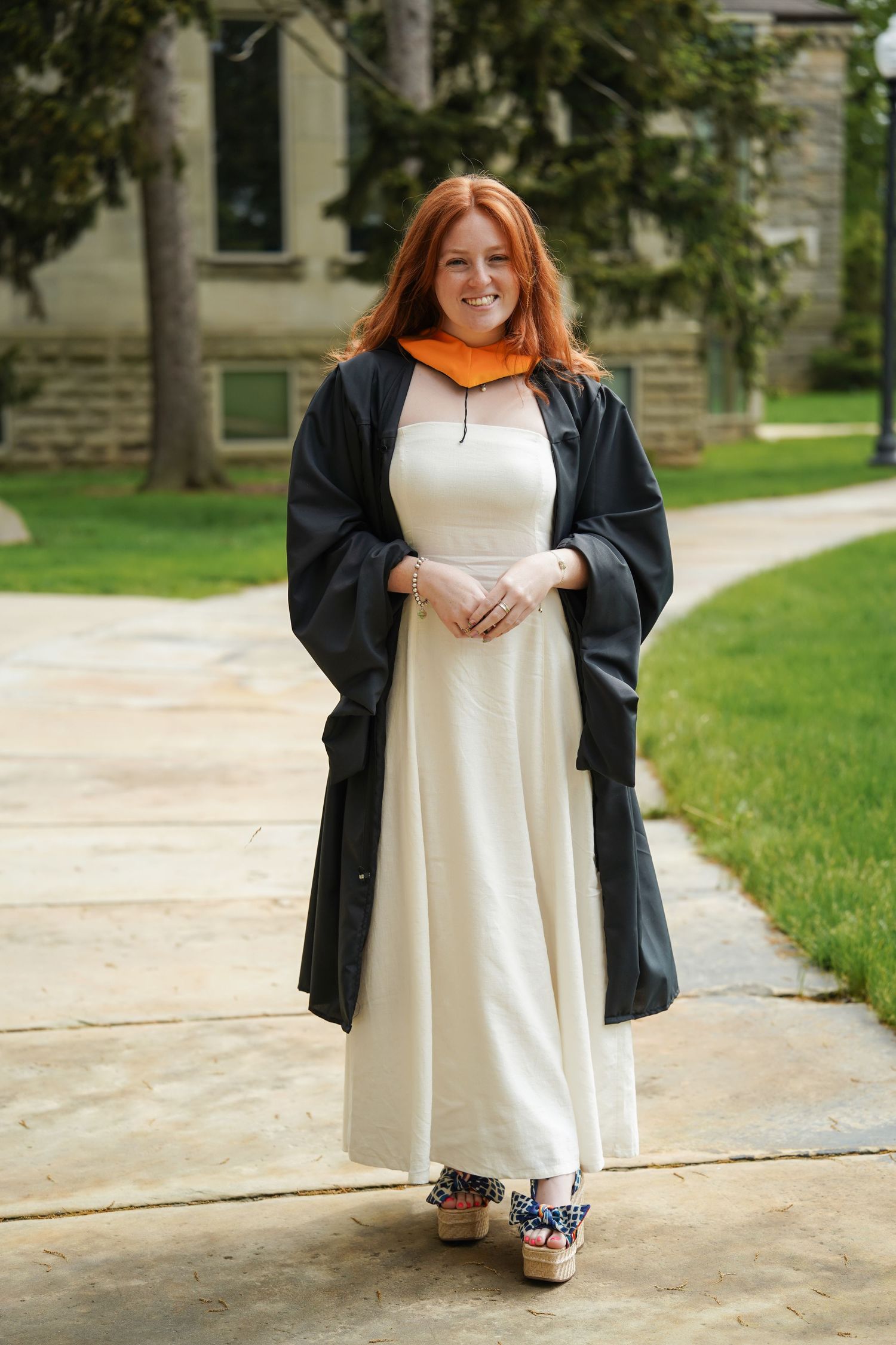 Graduate in black academic gown and white dress stands proudly on campus sidewalk during graduation day.