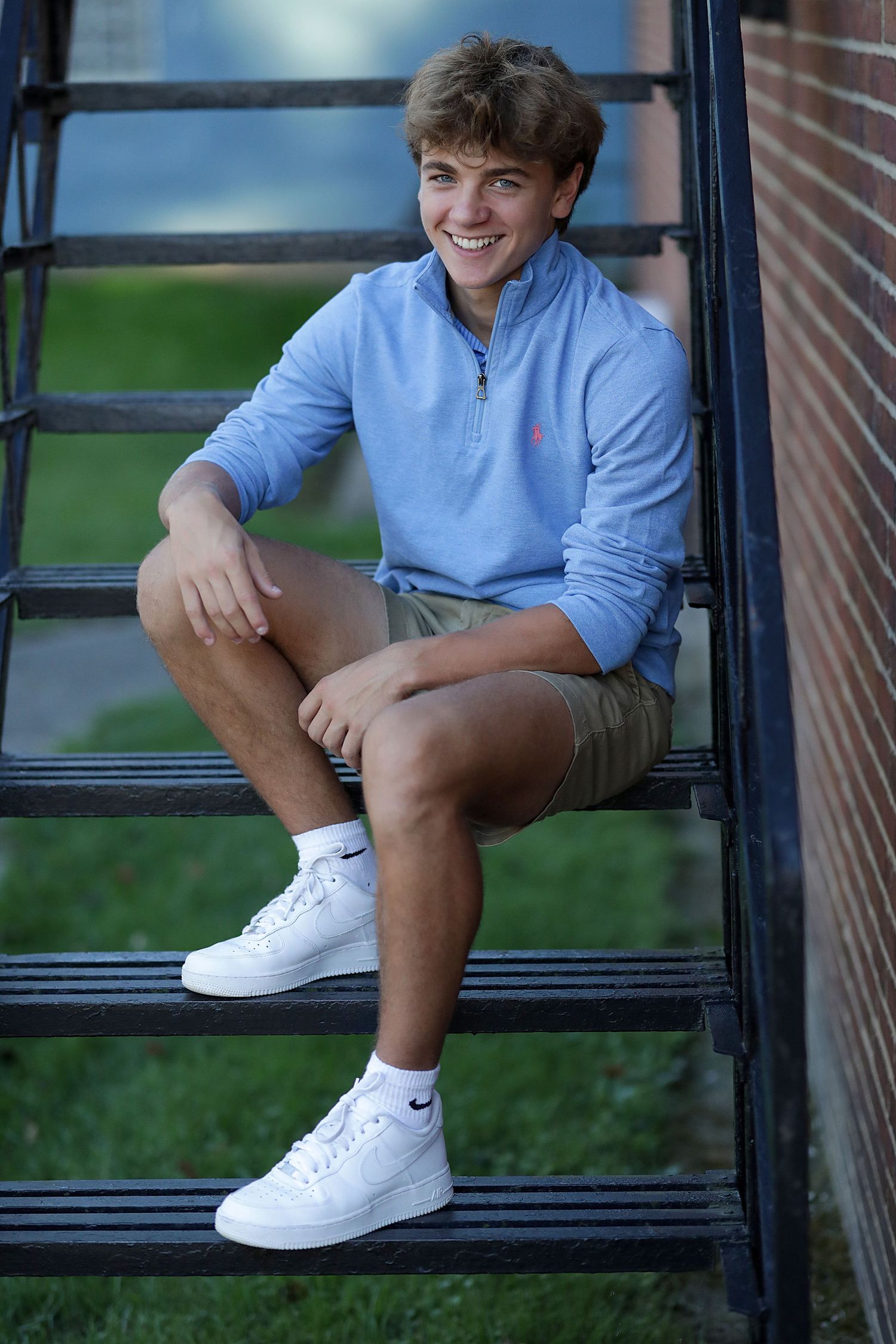 A person sits on outdoor black metal stairs wearing a light blue quarter-zip sweater, khaki shorts and white Nike sneakers.