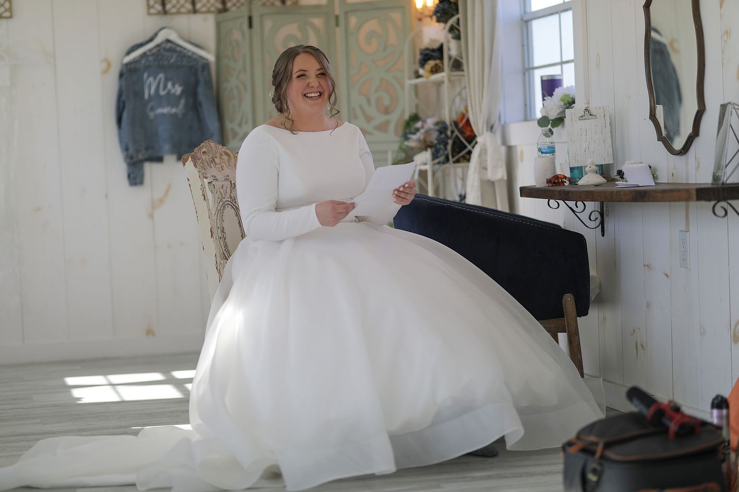 A bride in a long-sleeved white ball gown sits elegantly in a rustic white room.