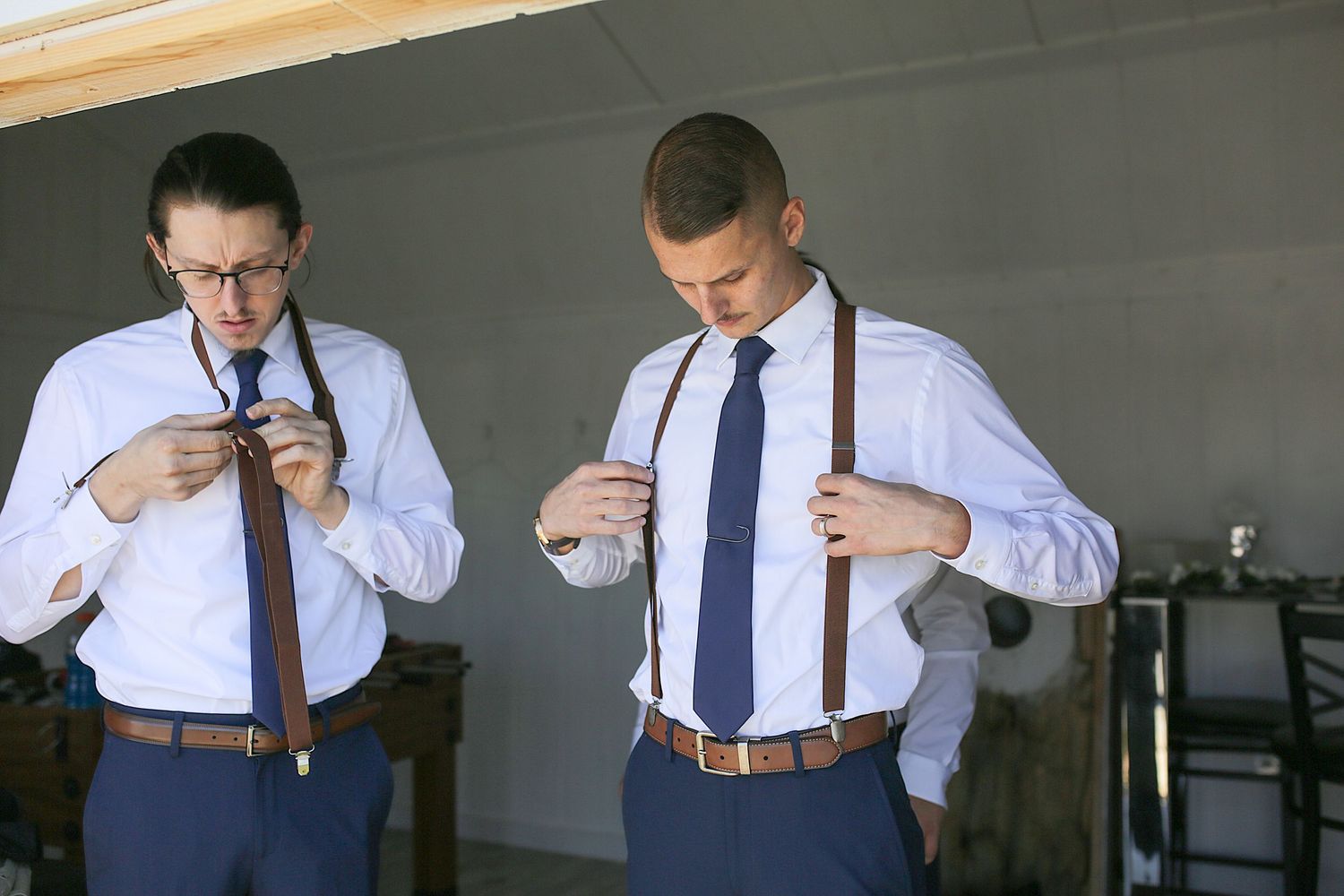 Two men in white dress shirts and navy ties adjust their leather suspenders while getting dressed.