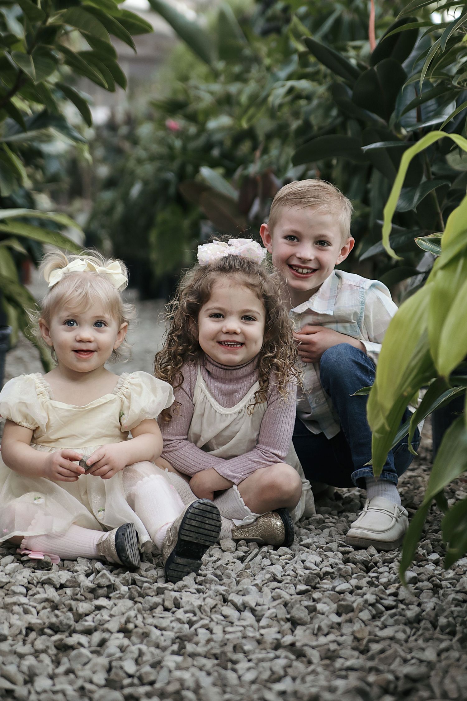 Children sit in a row together on gray gravel surrounded by lush green plants in a garden setting.