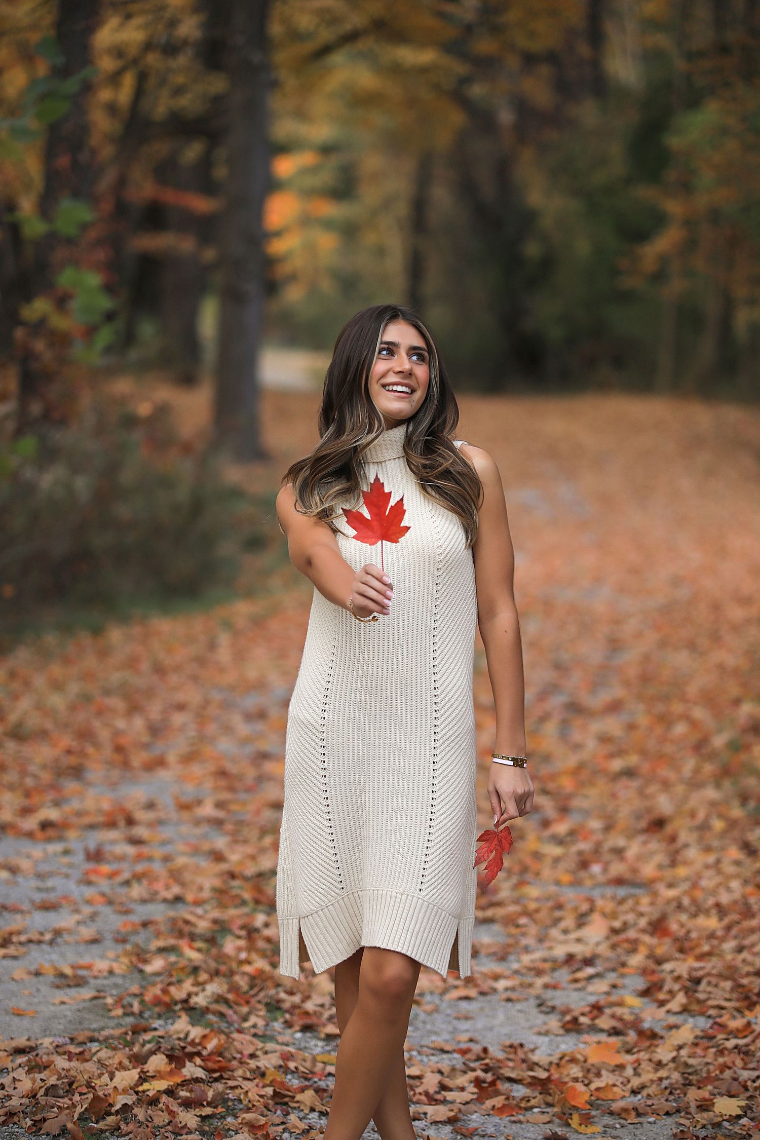 Someone in a white knit sweater dress poses playfully among fallen autumn leaves on a woodland path.