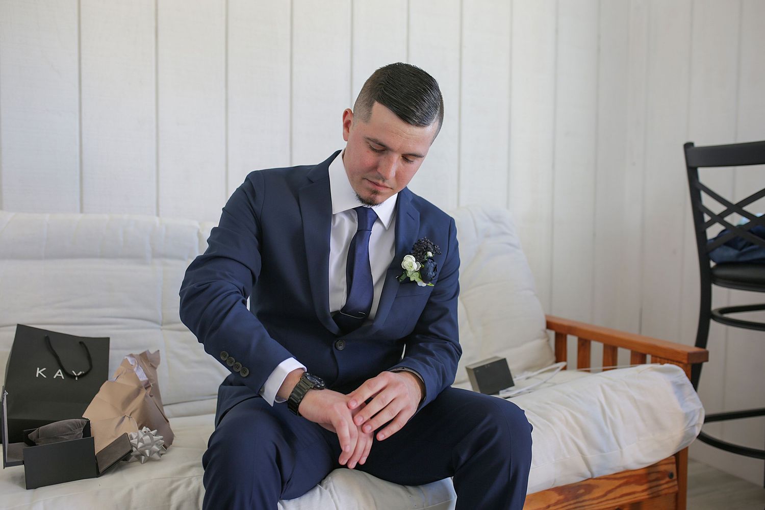 A groom in a navy blue suit and tie adjusts his cuffs while seated on a white bed.
