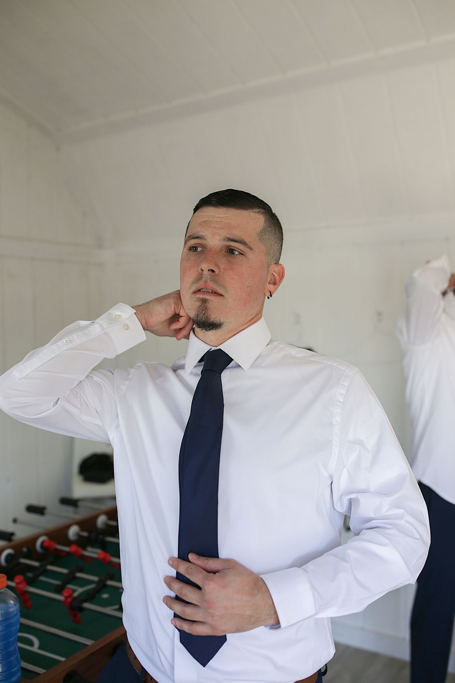 A wedding attendee adjusts his navy tie while getting ready in a white shirt.