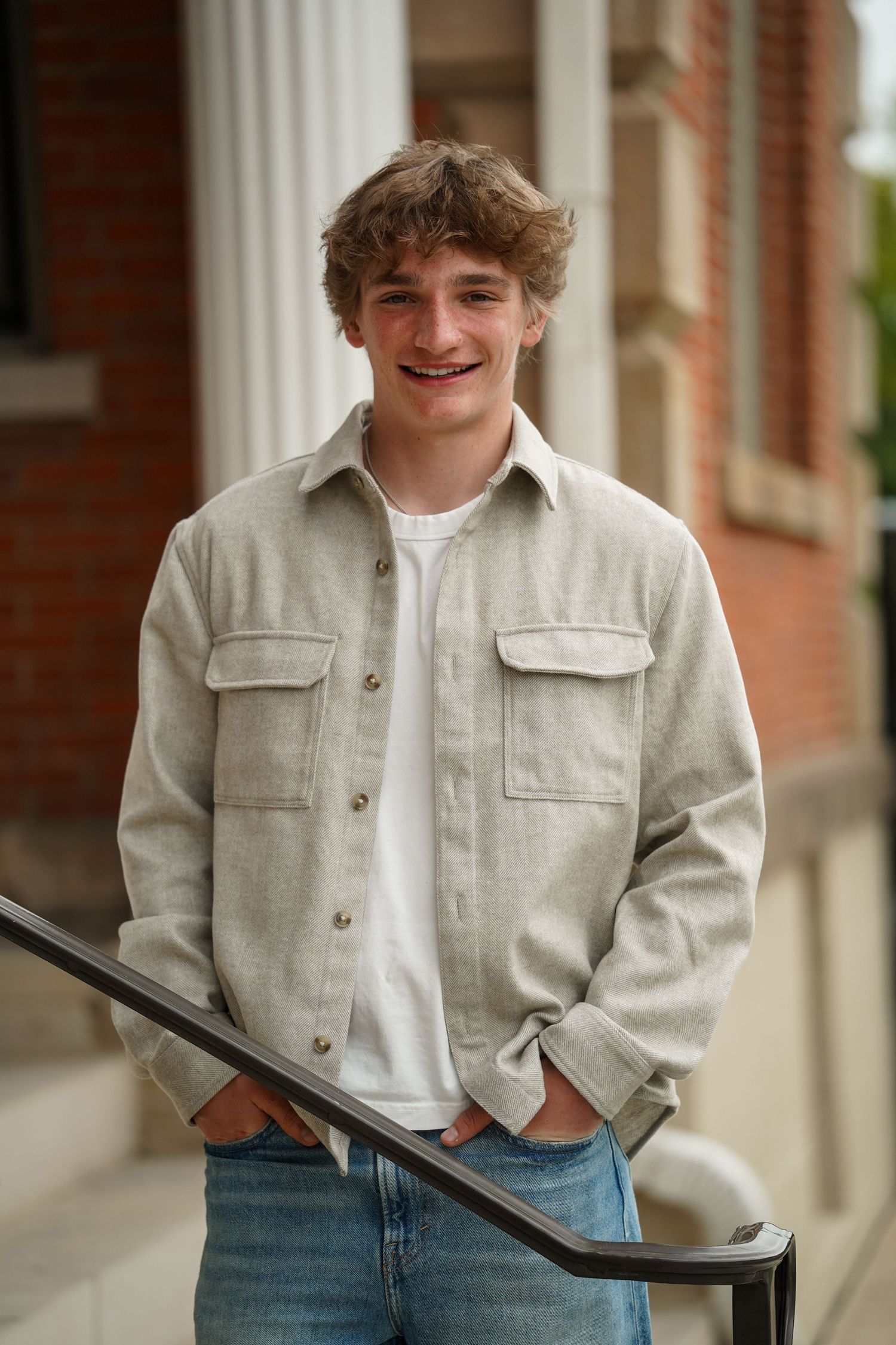 Young person in light jacket and jeans smiles and poses casually against brick building with columns.