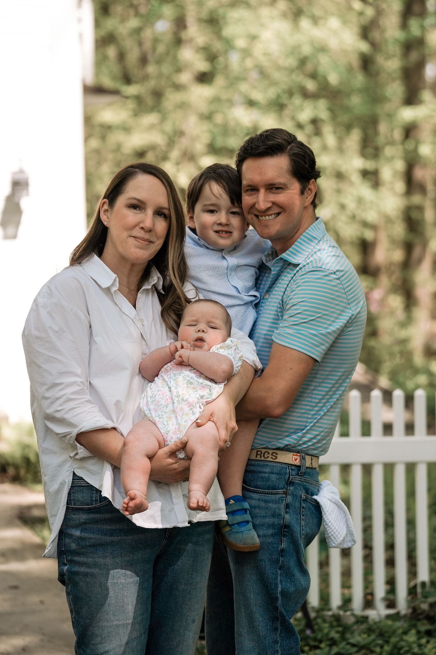 Happy family shares laughter and cuddles with baby in outdoor setting by white picket fence.