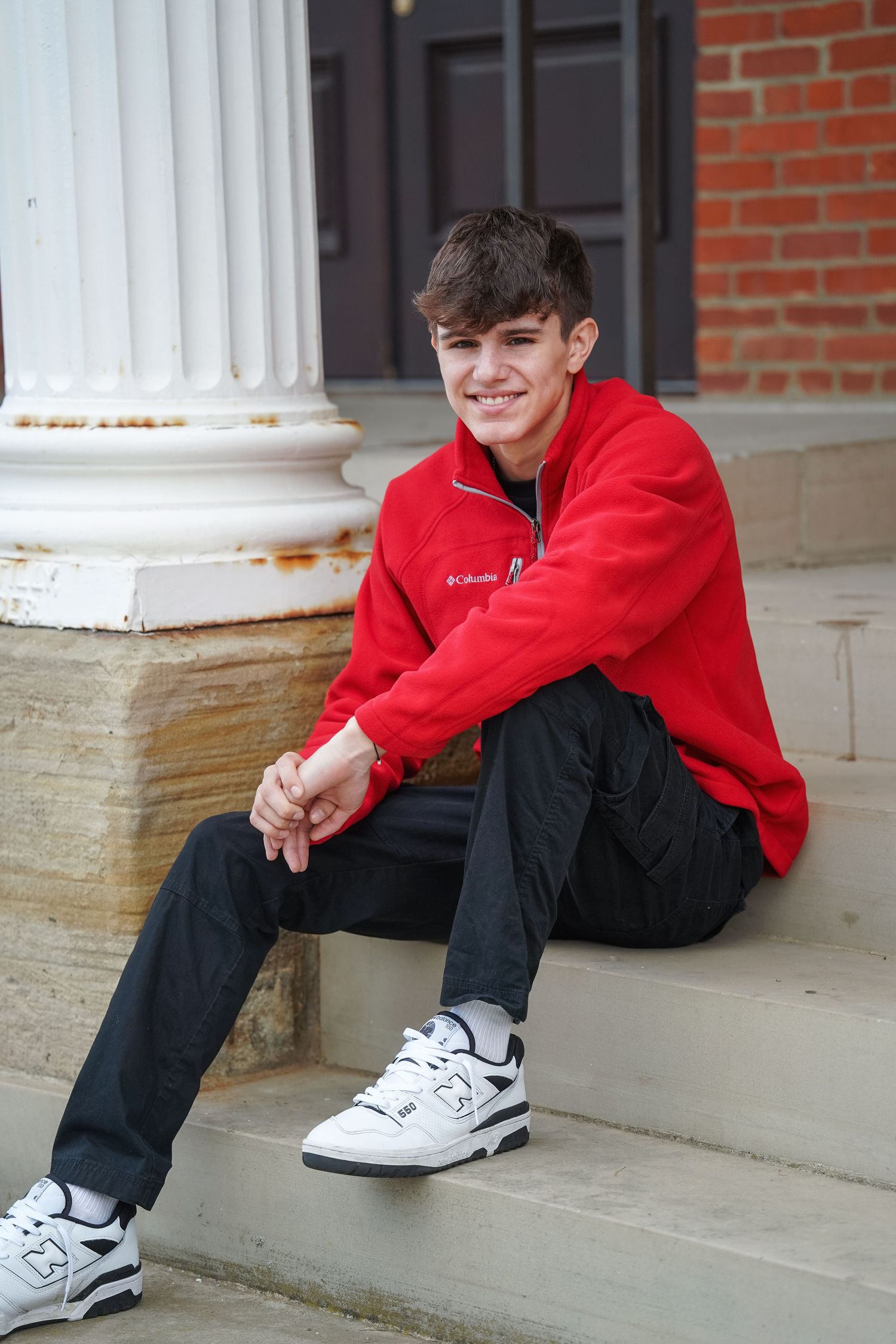 Person in a red sweatshirt and black pants sits on concrete steps wearing white athletic shoes.