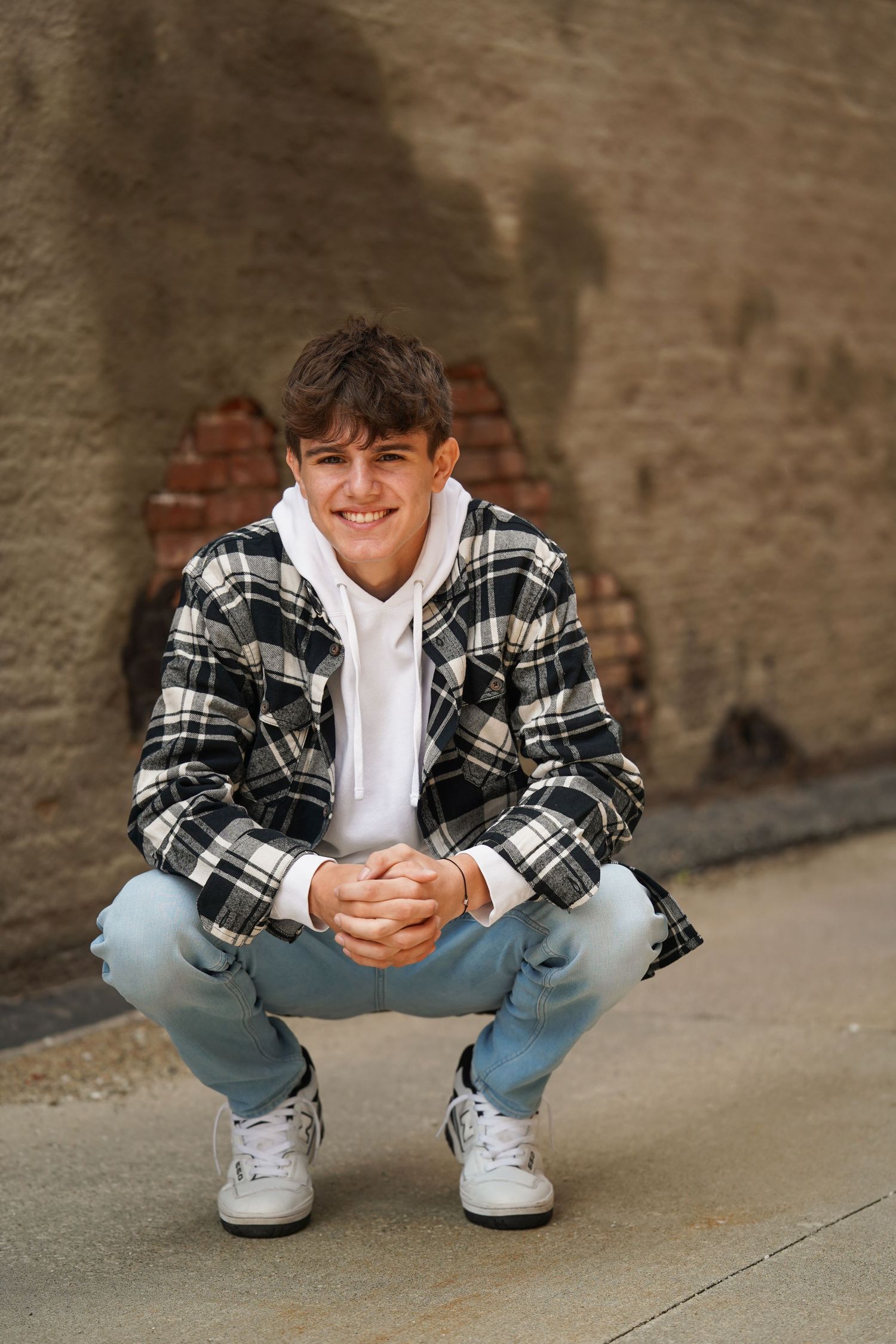 Young person in plaid flannel jacket and jeans crouches against brick wall wearing white hoodie and sneakers.