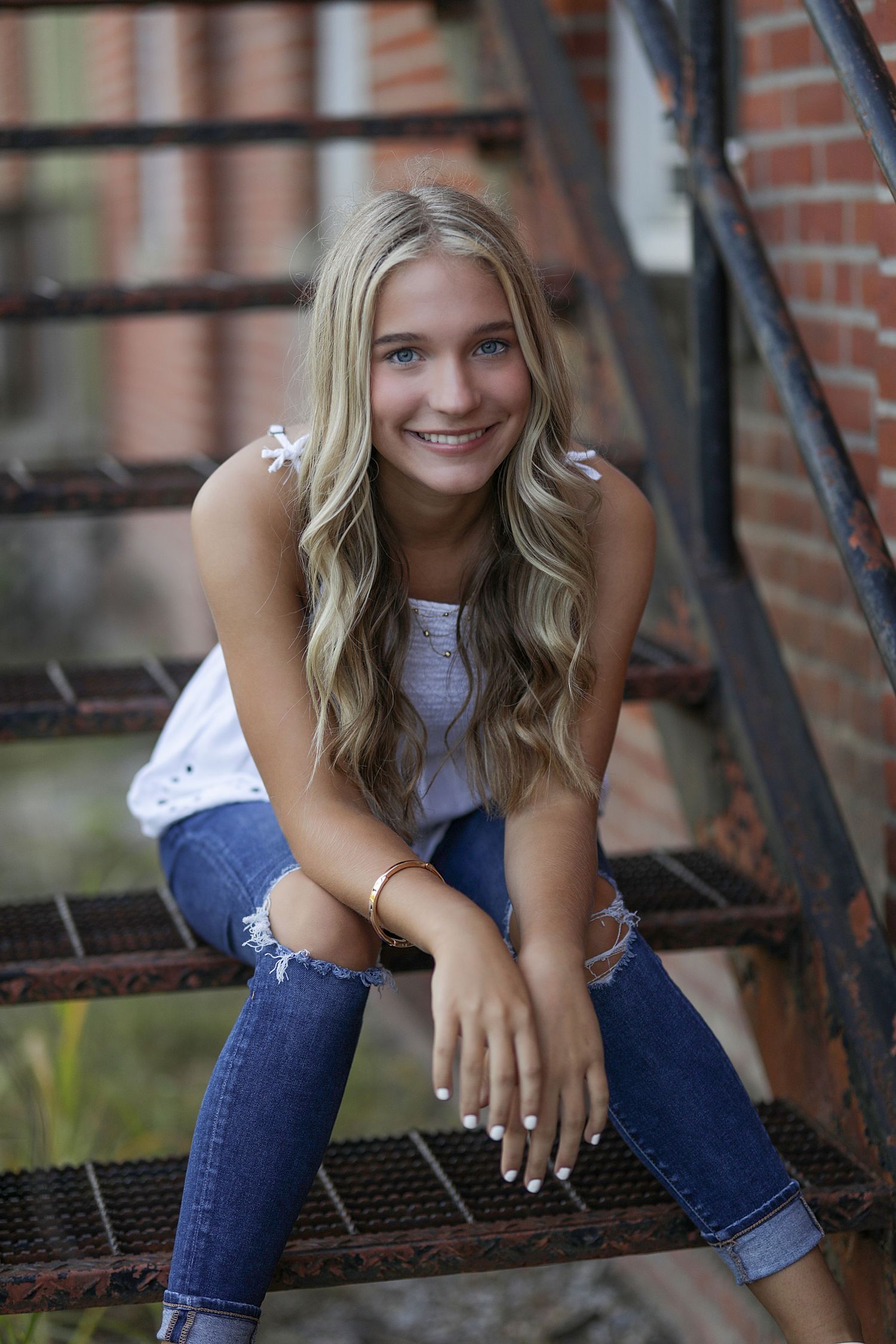 Smiling young woman in white top and ripped jeans sitting casually on metal stairs with brick wall background.