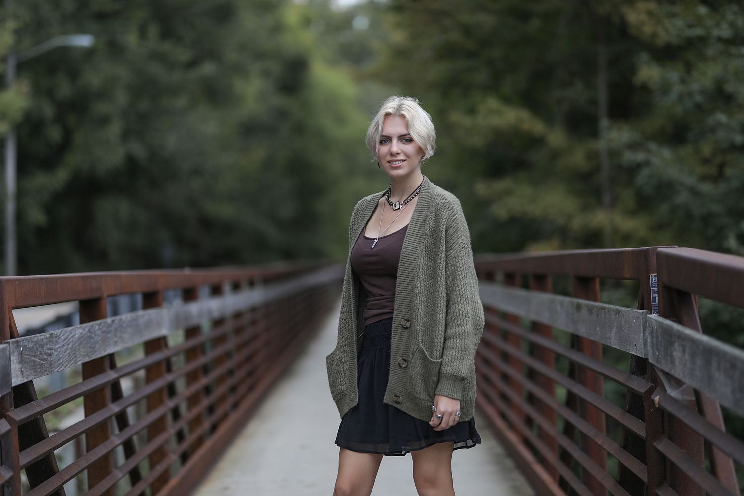 A person wearing a green cardigan and black dress stands on a wooden bridge pathway in a forested area.