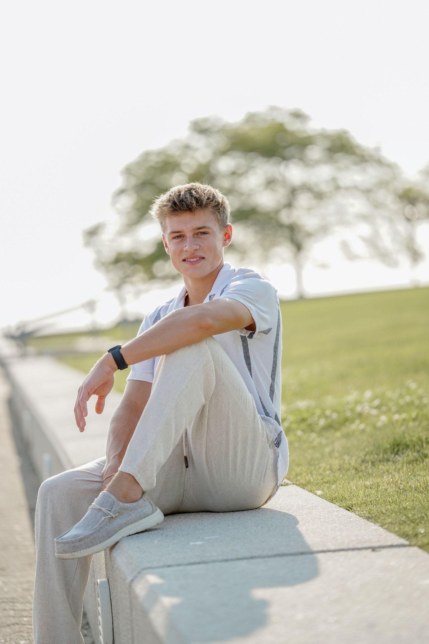A young person in a light blue shirt and white pants sits casually on a concrete barrier outdoors on a sunny day.
