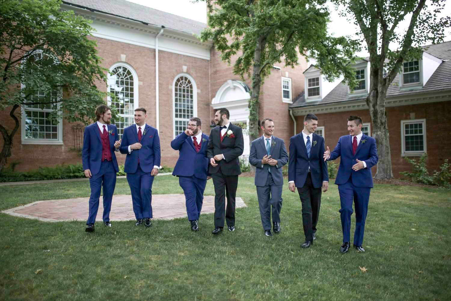 A group of people wearing navy blue suits stands in formation on a green lawn near brick buildings.