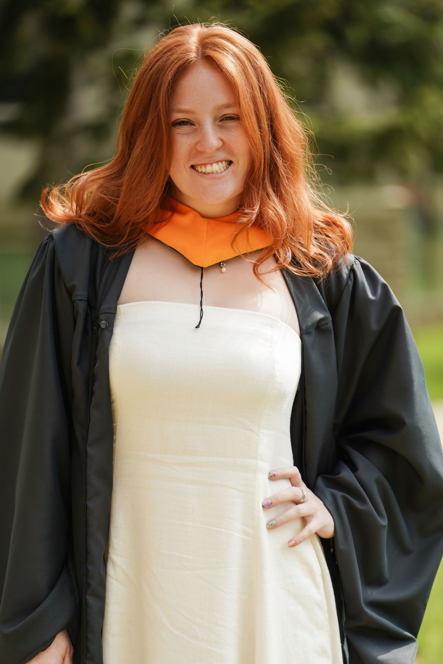 Graduate wearing black graduation gown over white dress stands outdoors with orange highlights against blurred background.