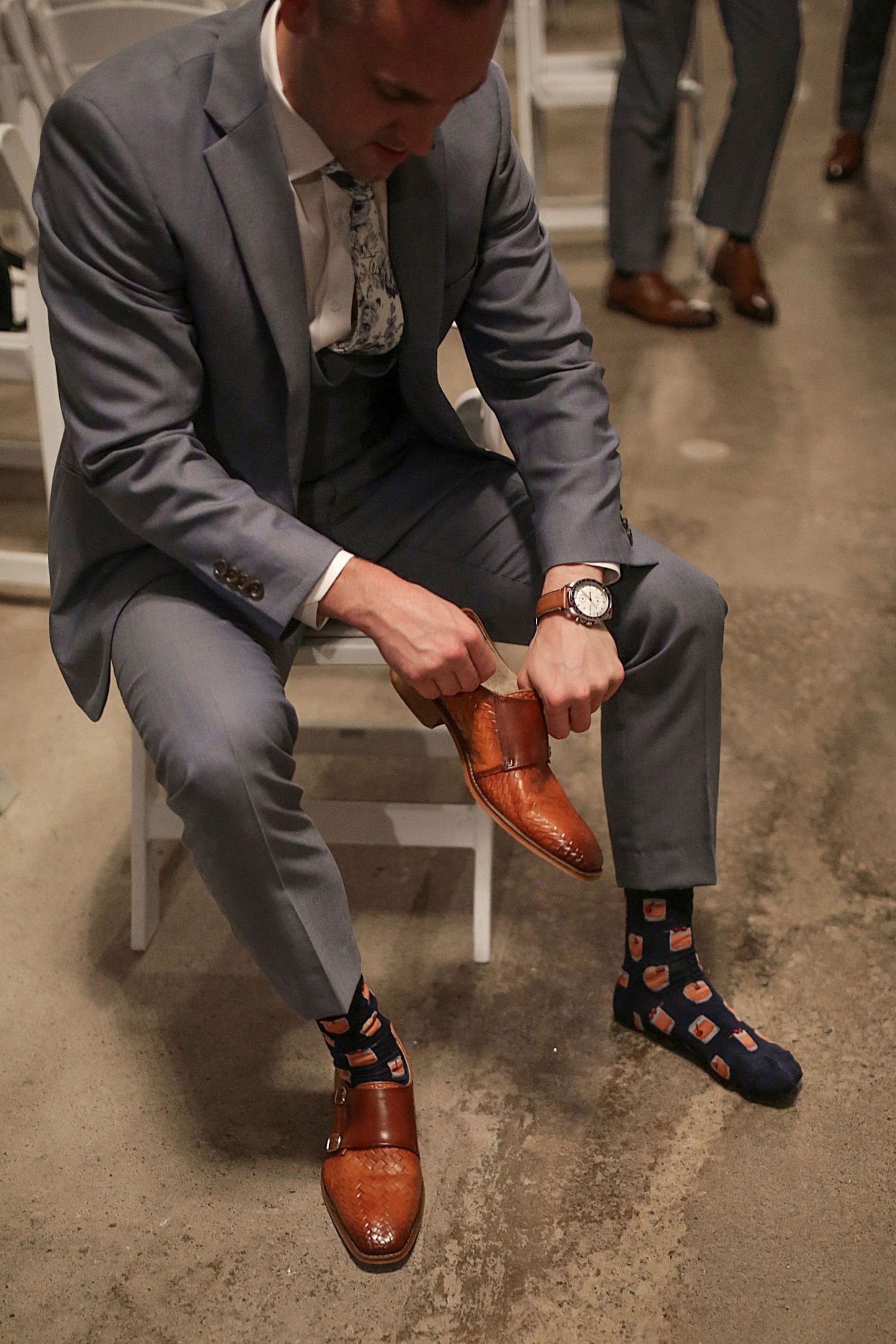 Groomsman in grey suit puts on brown leather dress shoes while showing colorful socks.