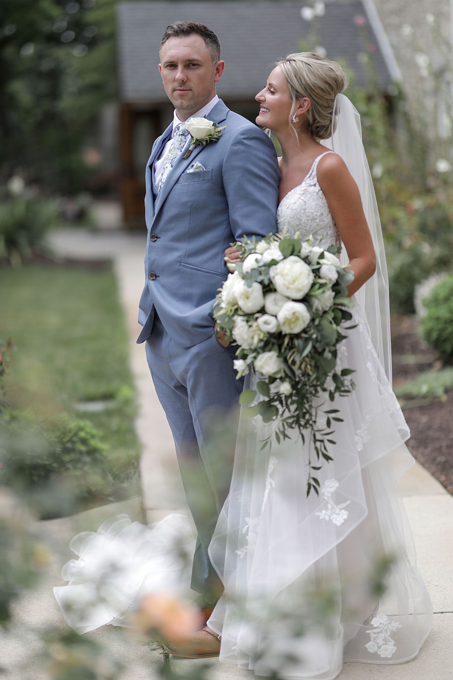 Wedding couple poses in garden setting with white floral bouquet and gray suit.