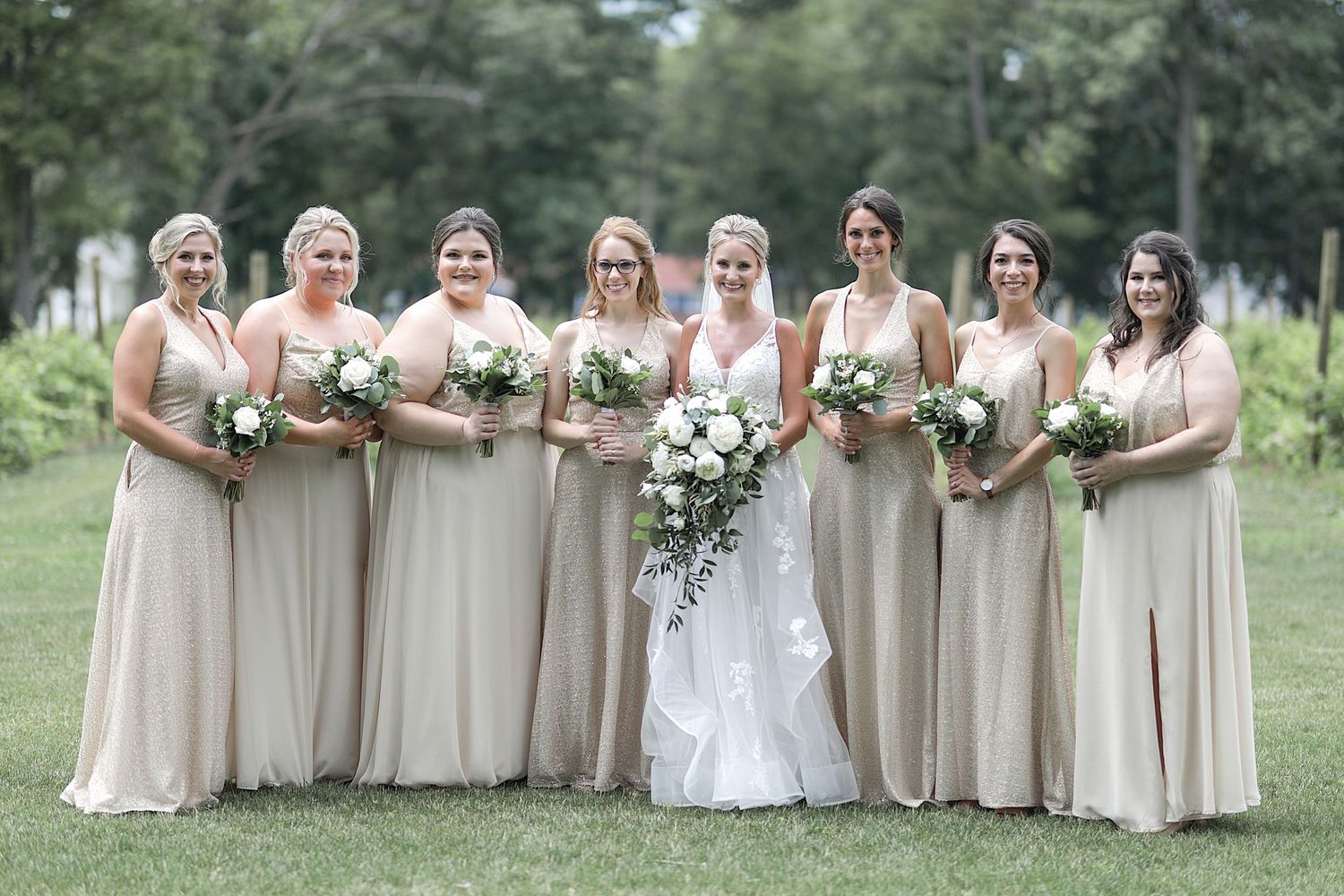 Wedding party in cream colored dresses poses with bride in white gown outdoors.