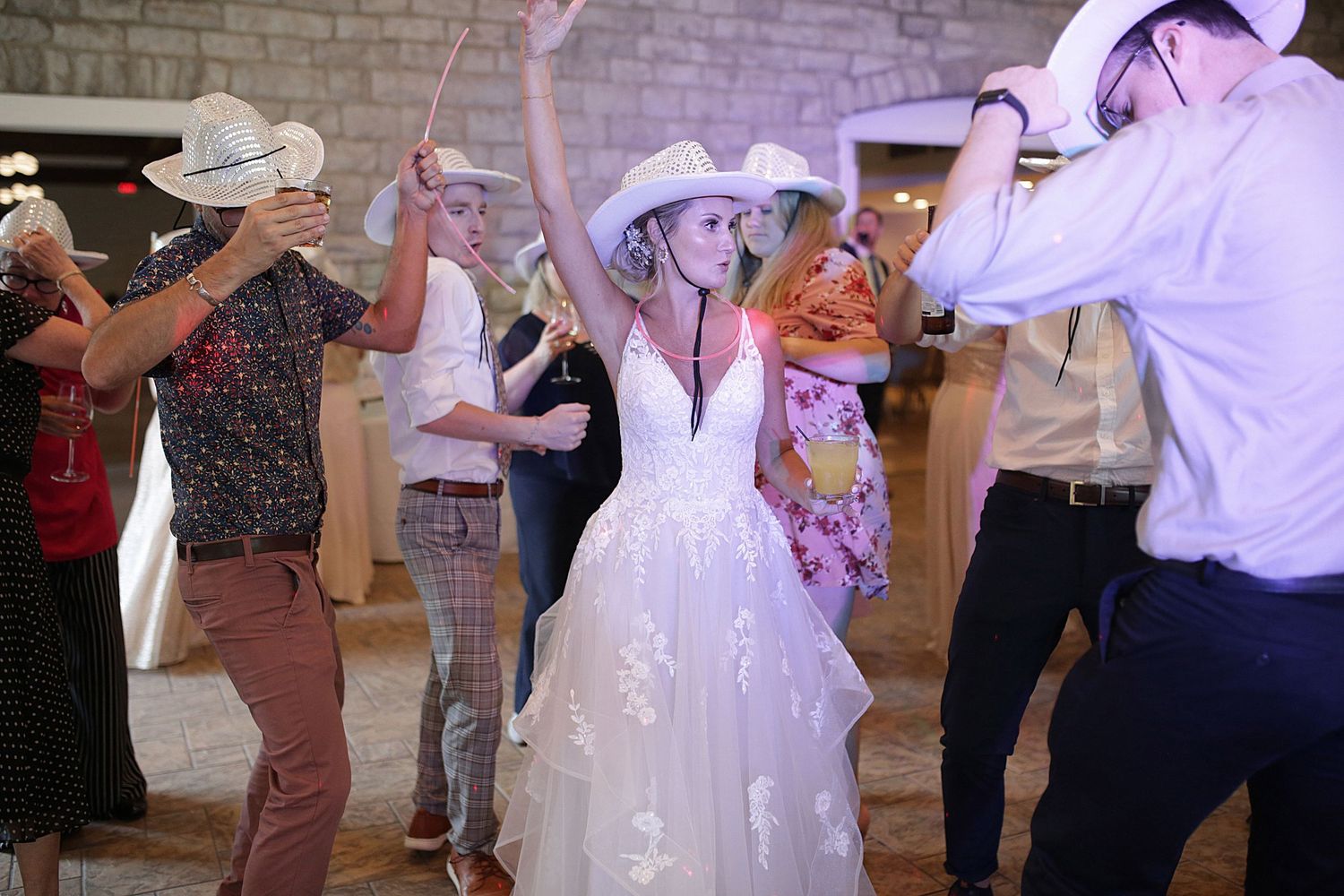 Wedding reception dance party with guests wearing cowboy hats in a rustic venue.