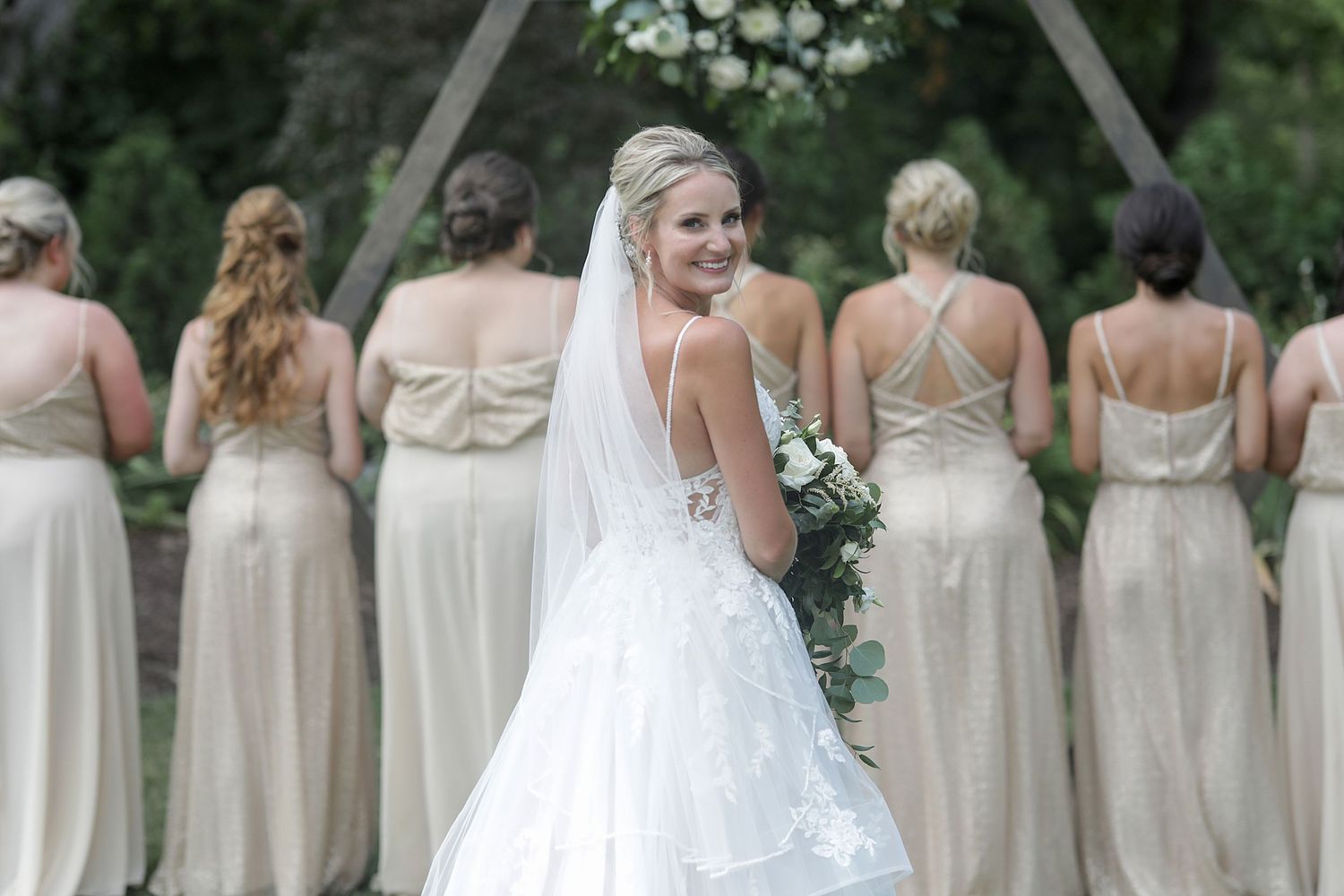 Row of bridesmaids in champagne dresses stands outdoors in a garden setting. Cleveland wedding photographer