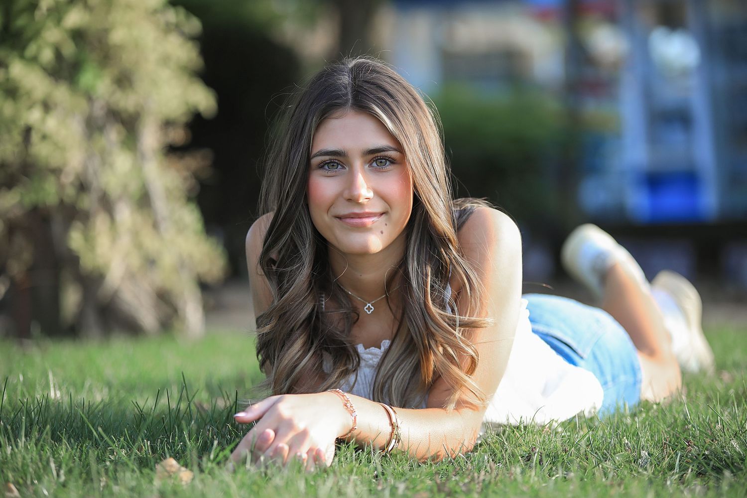 A person with wavy brown hair lies casually on green grass during a sunny outdoor portrait session.