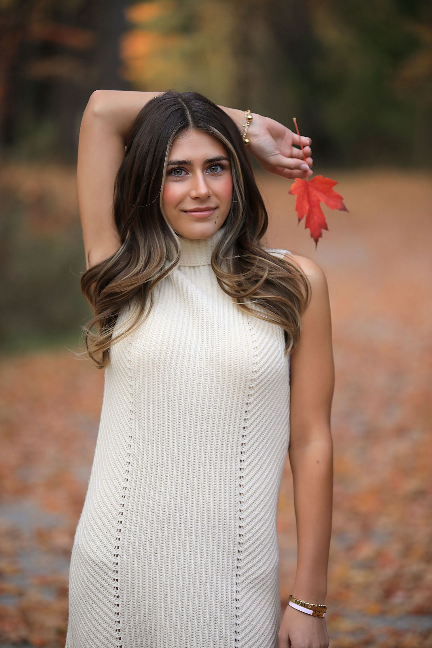 Person in cream sweater dress holds red autumn leaf while posing outdoors with fallen leaves in background.