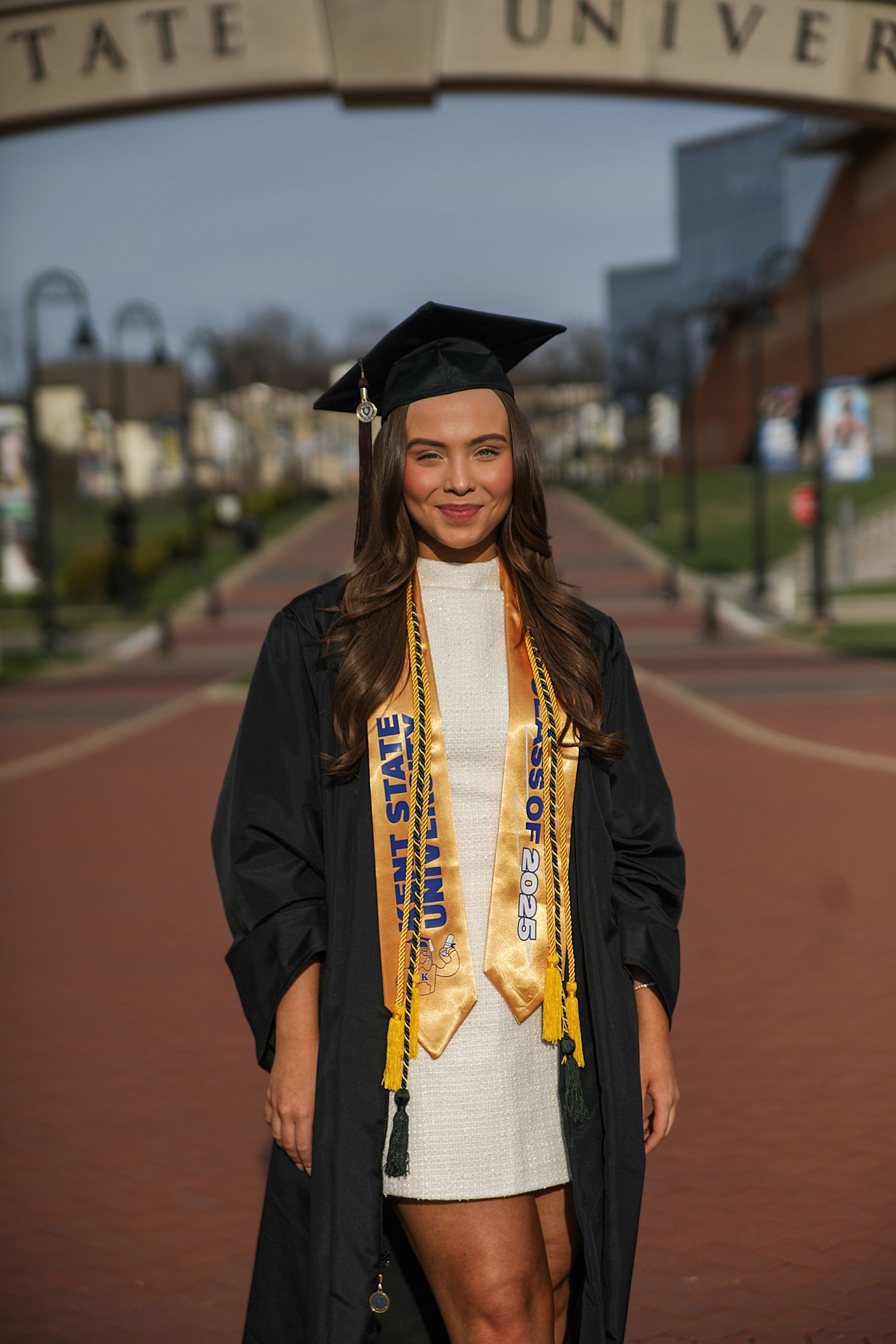 Graduate in cap and gown poses outdoors wearing gold stole and graduation regalia on a brick walkway.