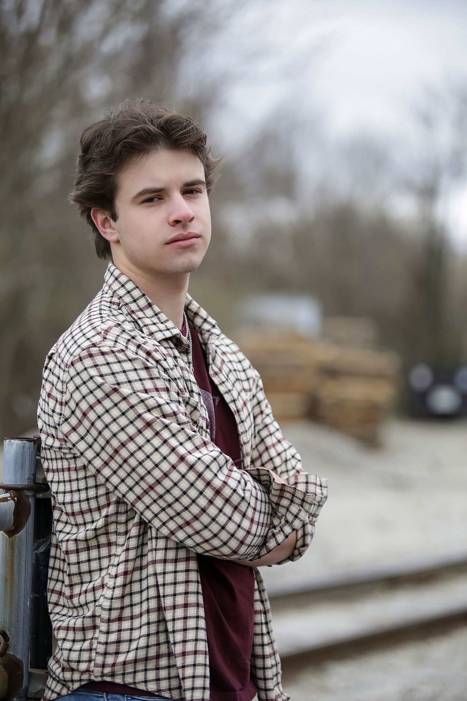 A person in a checkered shirt and burgundy top poses thoughtfully against a blurred outdoor background.