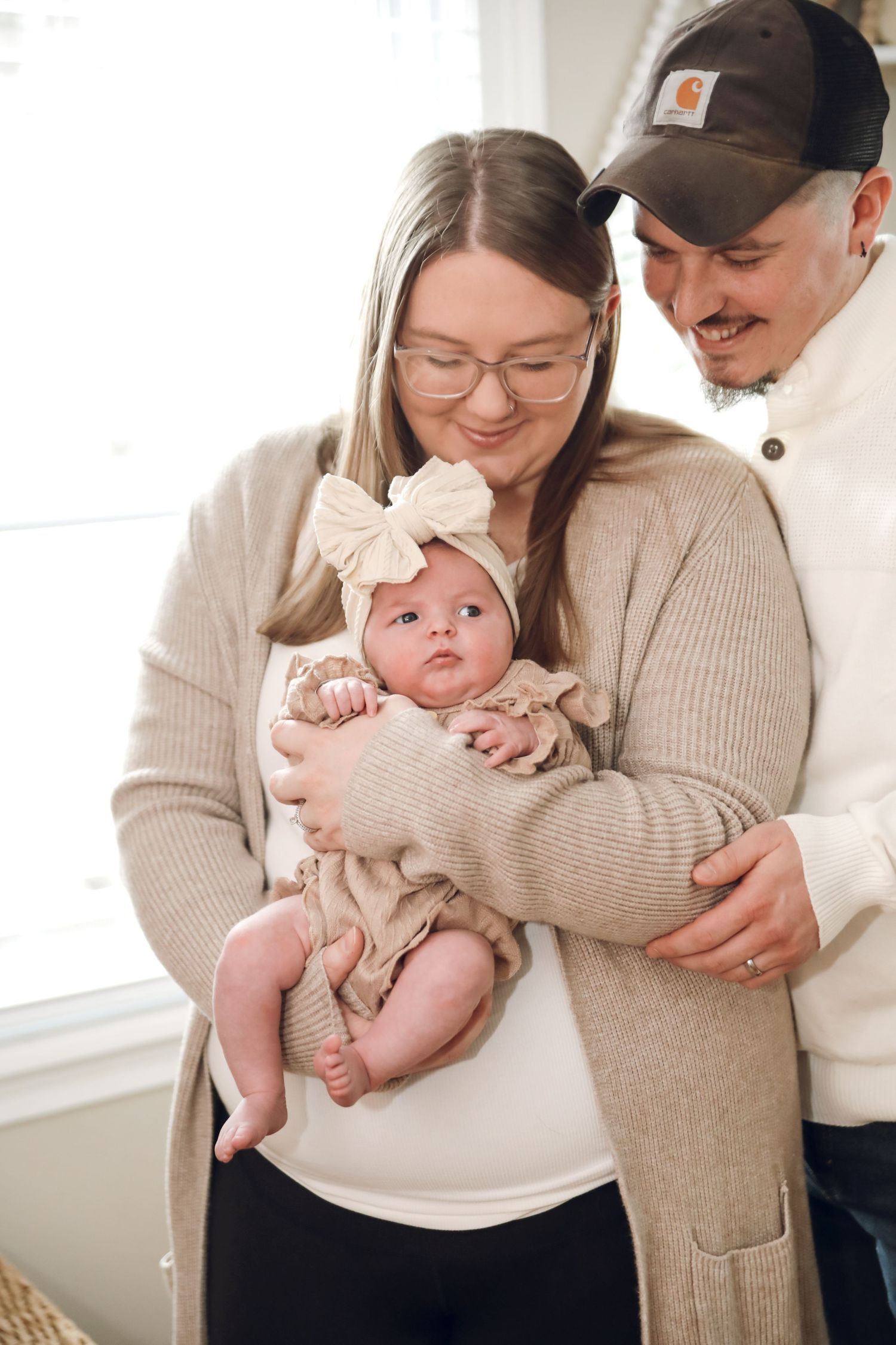 A collage of tender moments showing parents in beige sweaters holding and cuddling their baby in a bright room.