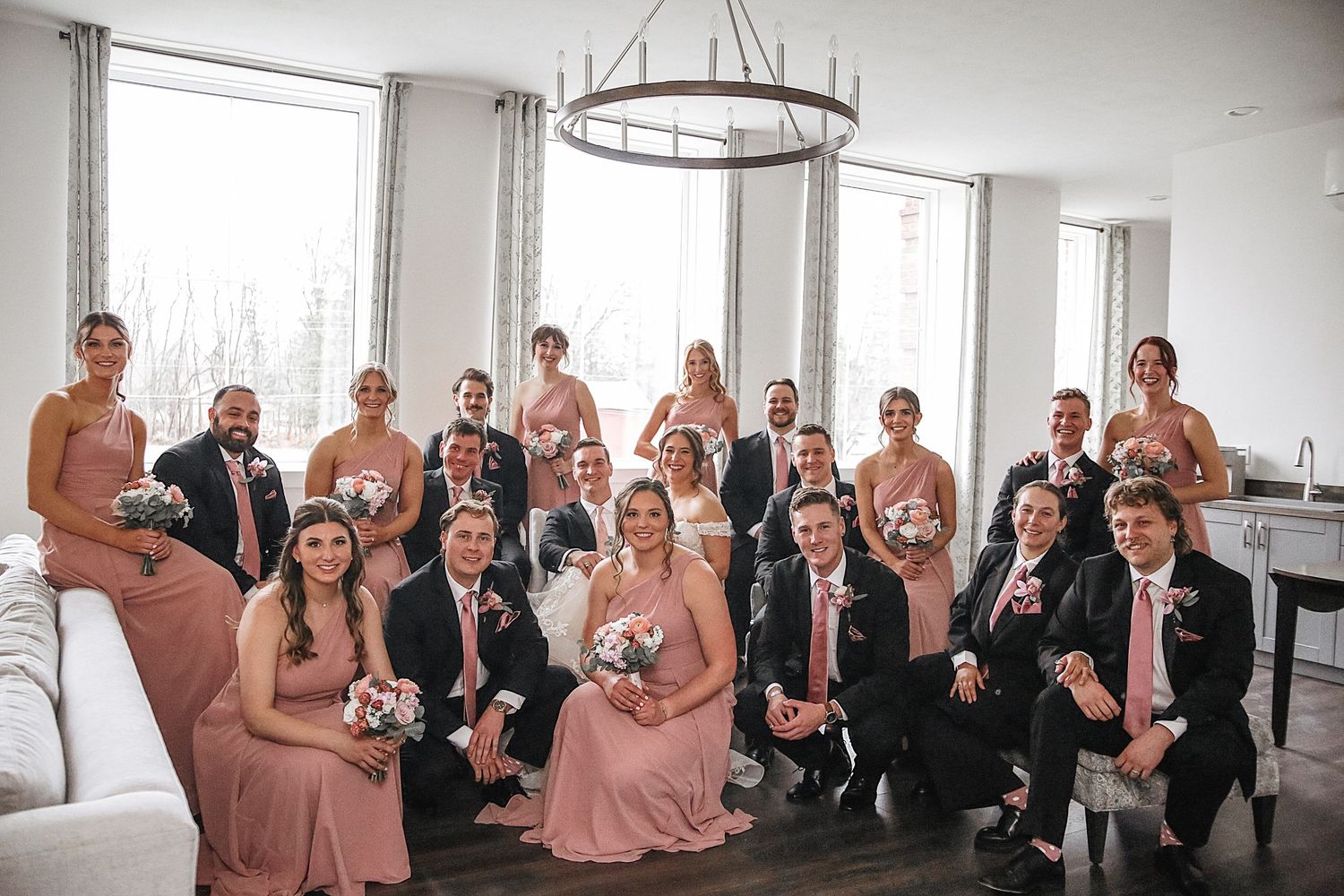 Wedding party in formal attire poses for candid photo series in bright white room with chandeliers.