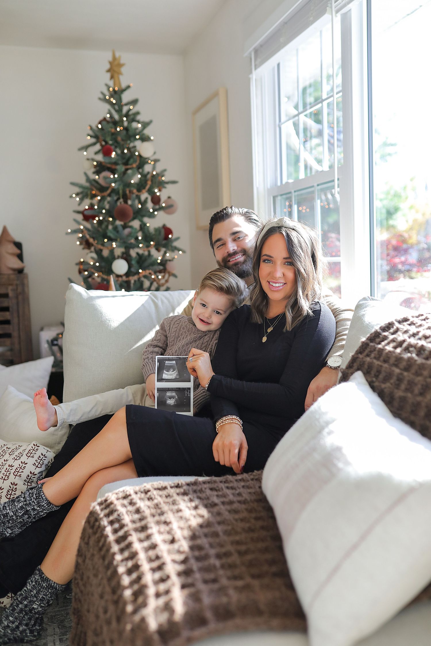 Family sits on couch together holding ultrasound photo with decorated Christmas tree in background.