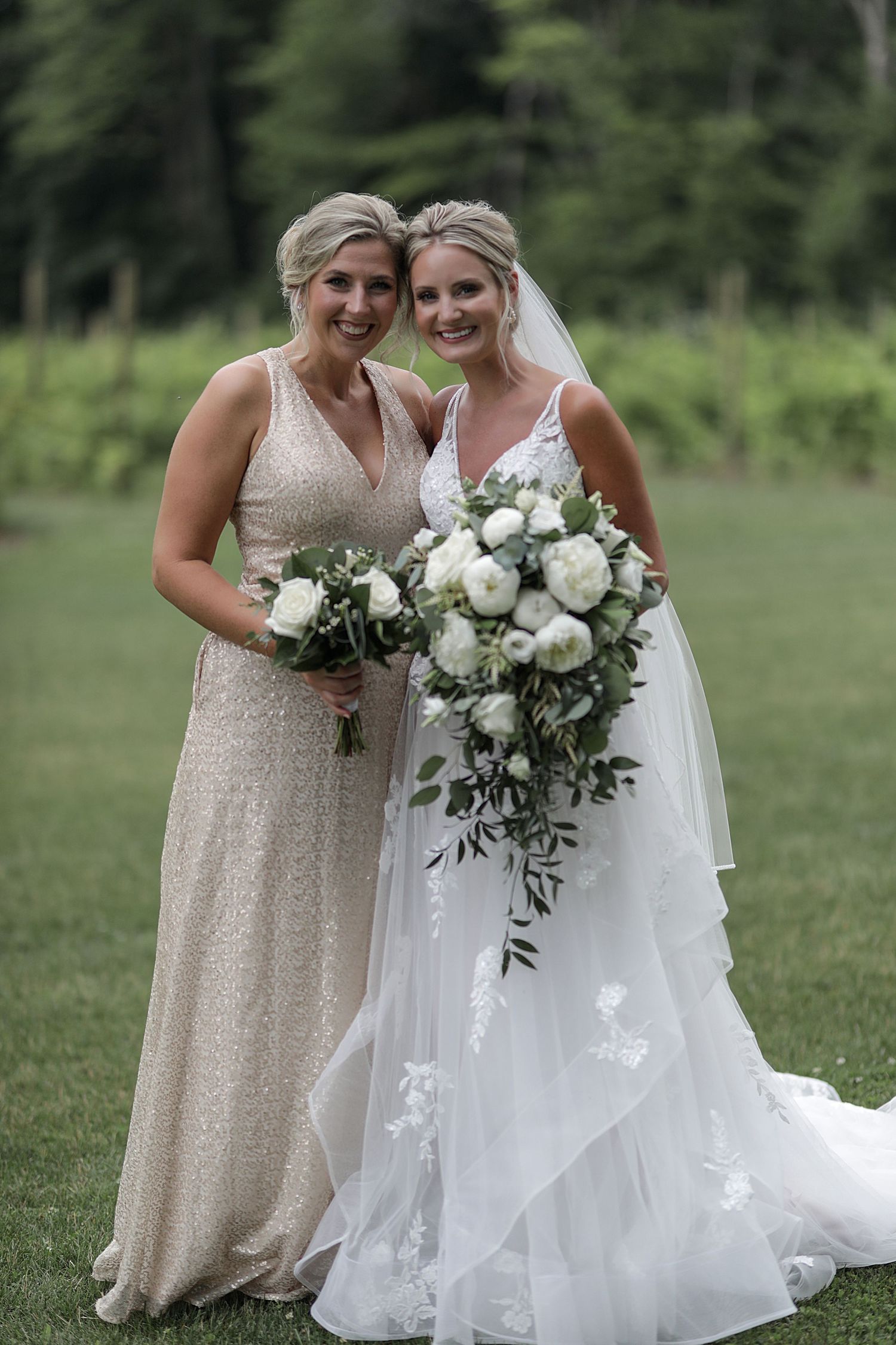 Wedding portrait of two women in formal dresses holding white bouquets against a green outdoor backdrop.
