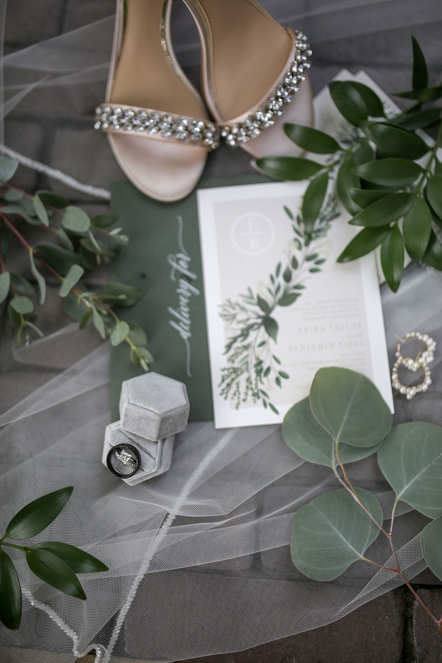 Wedding details flat lay with crystal shoes, eucalyptus leaves, invitation and ring box on veil.
