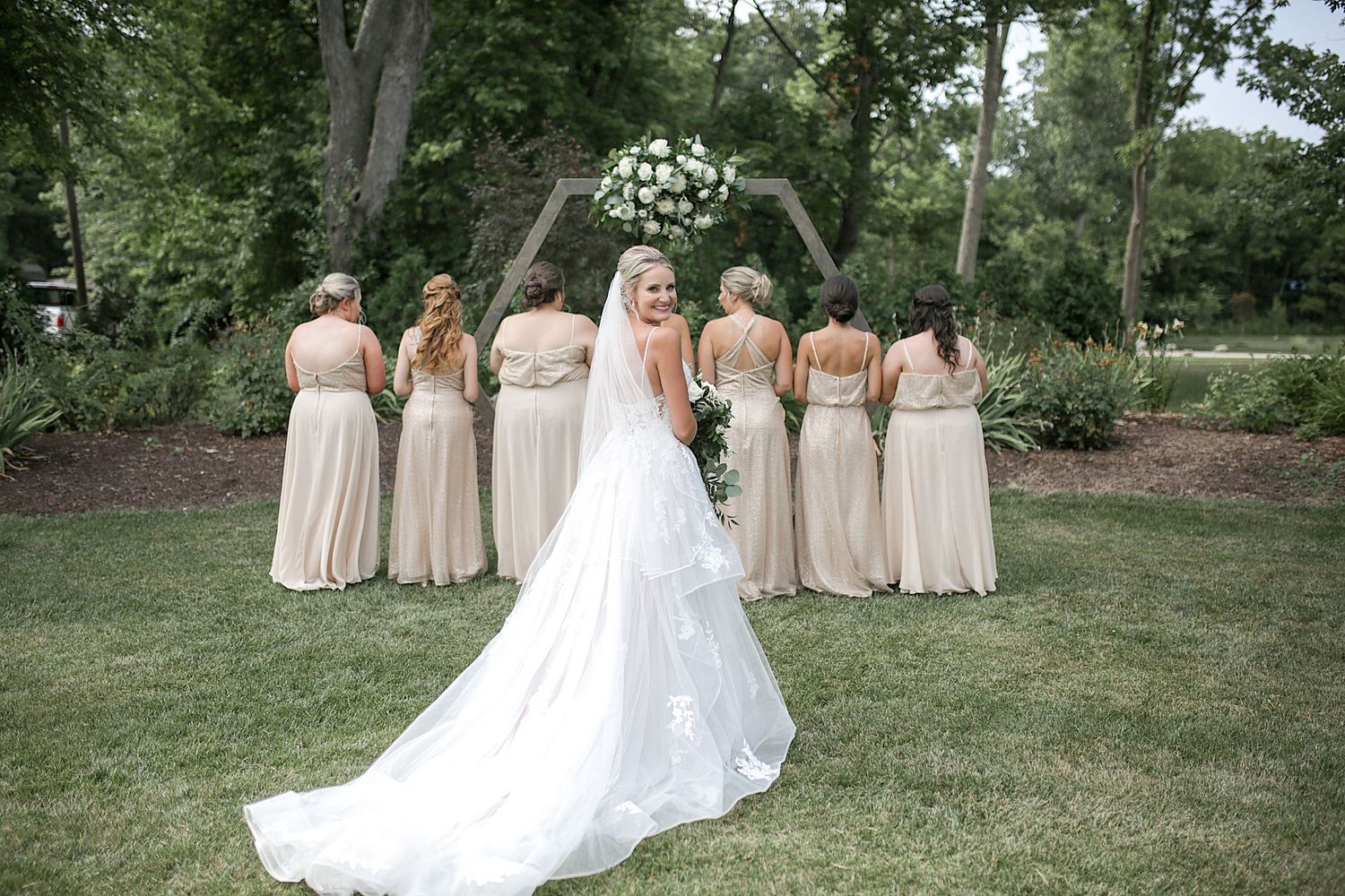 Wedding party poses in cream dresses and white gown against green garden backdrop.