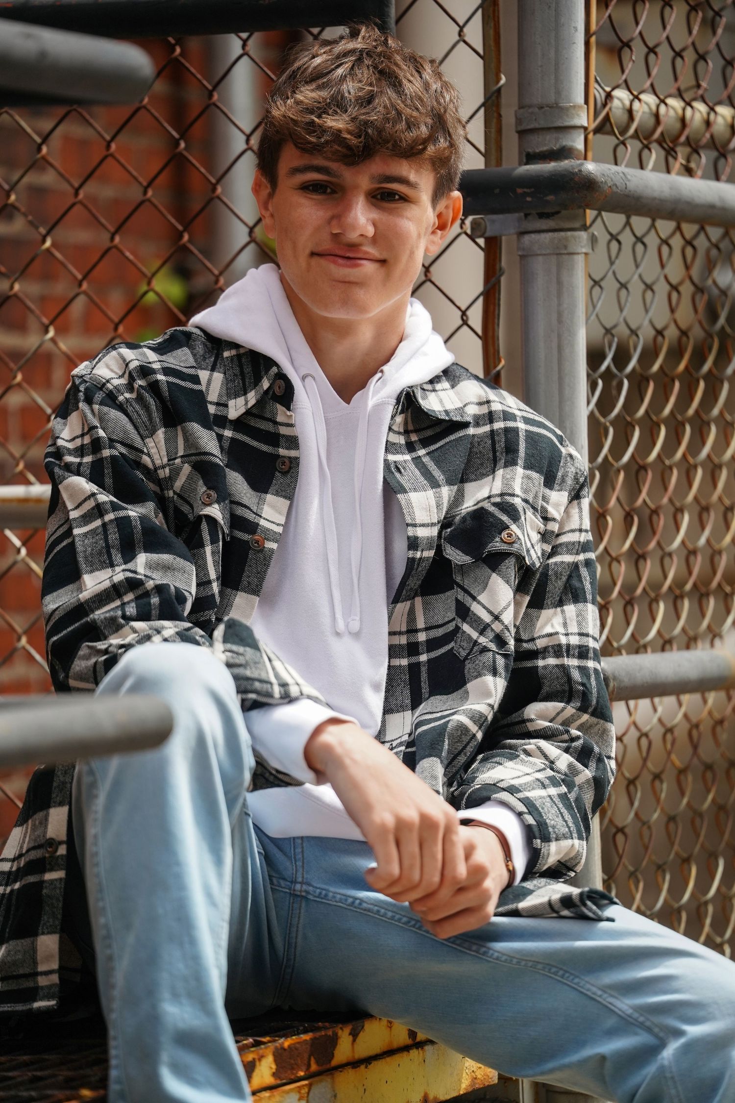 A young person in a plaid flannel shirt and jeans sits casually against a chain link fence.