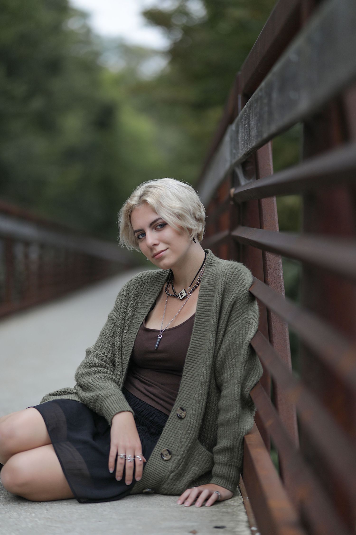 Portrait of a short-haired person wearing an olive cardigan and tank top sitting on a wooden bridge in soft lighting.