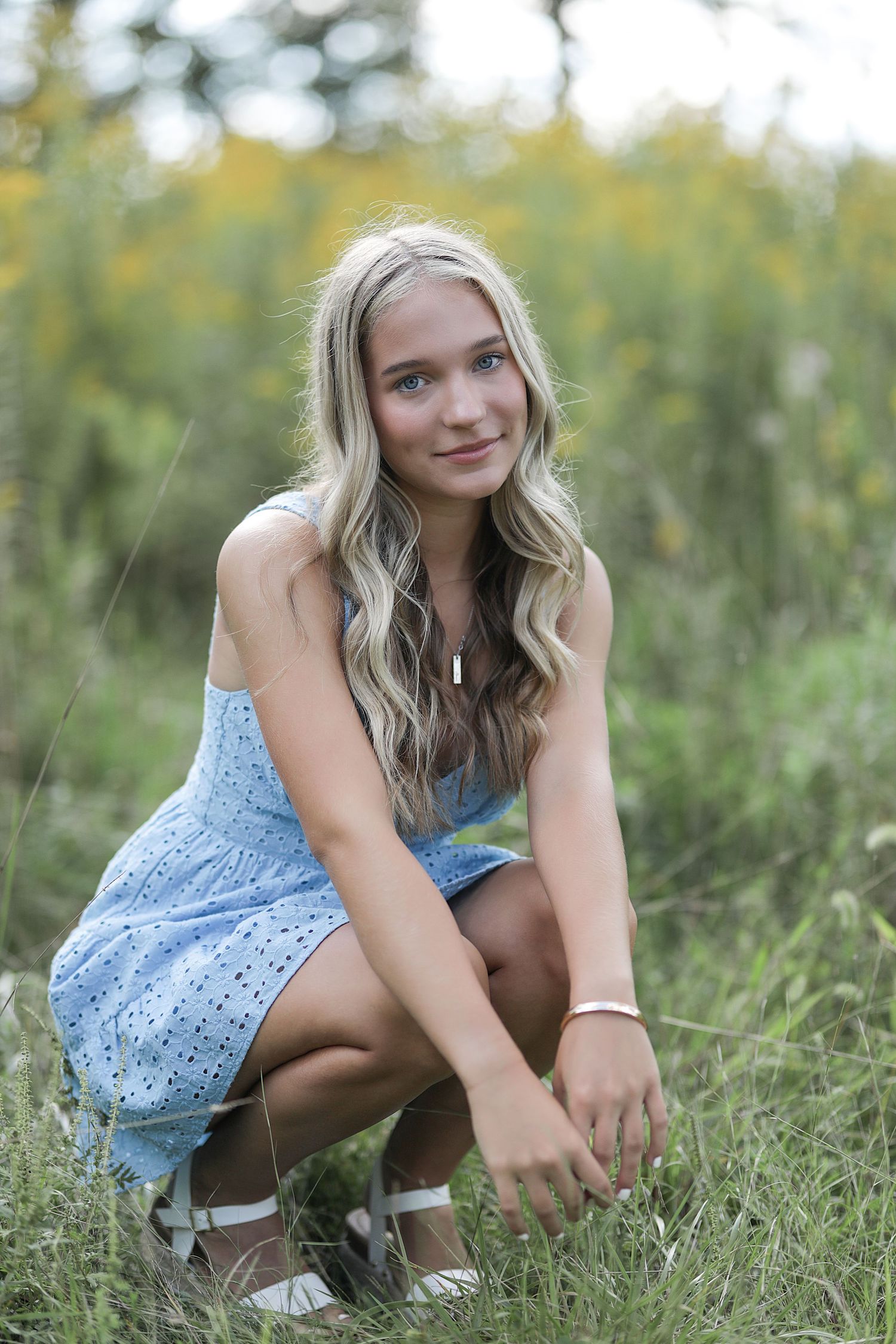 A young woman in a light blue lace dress crouches in a grassy field with wildflowers in the background on a sunny day.