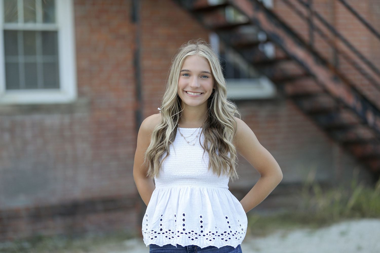 A woman in a white eyelet lace top stands outdoors near a brick building with fire escape stairs.