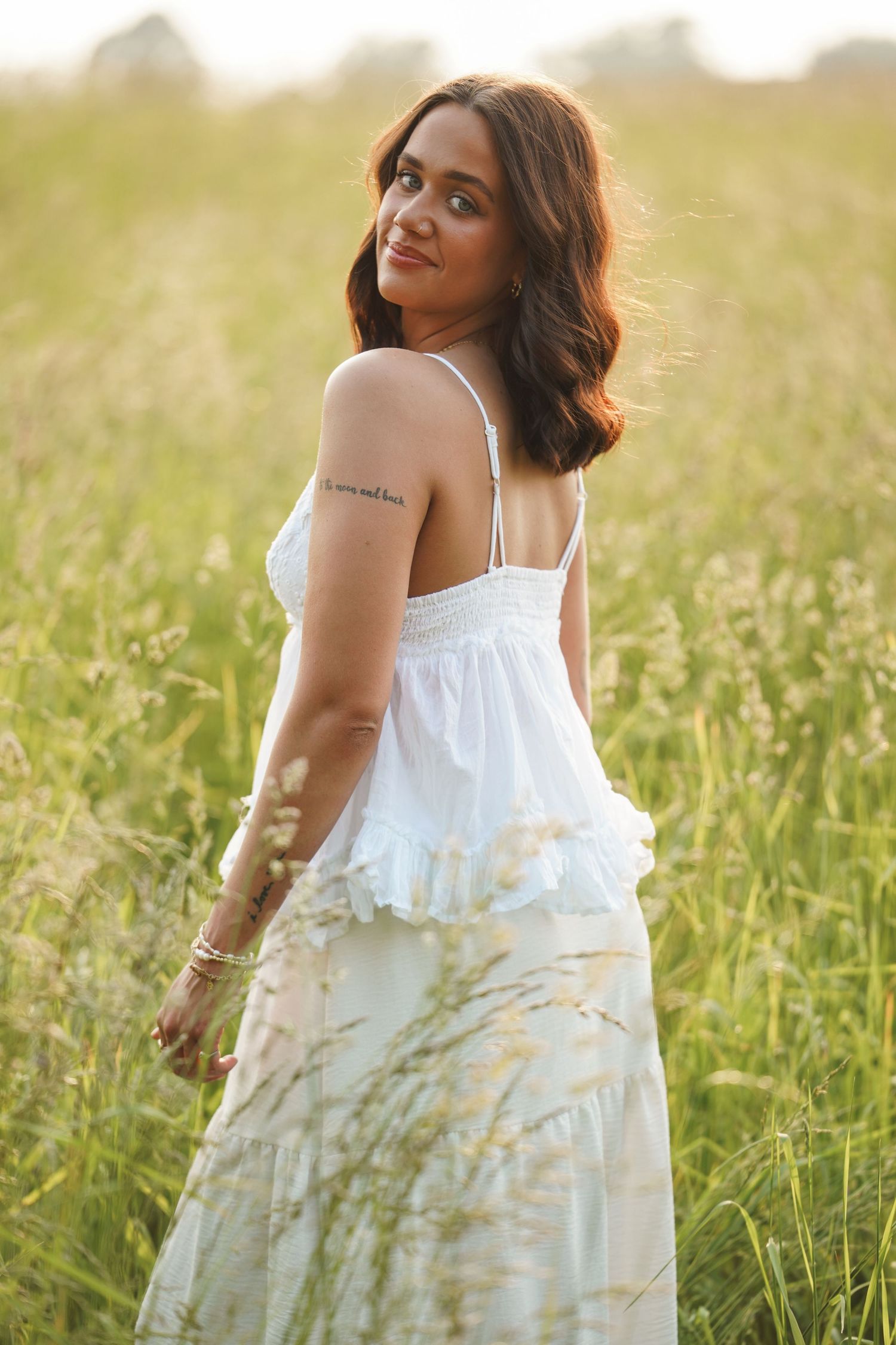 A woman in a flowing white sundress poses gracefully in a sunlit field of tall grass during golden hour.