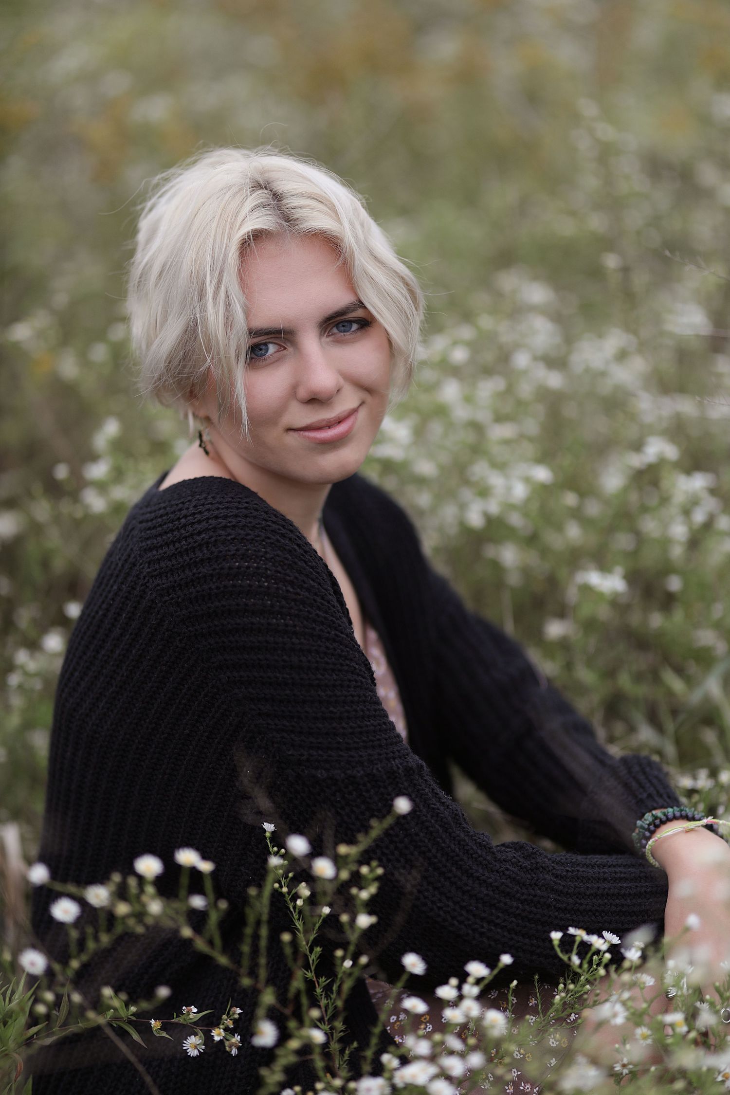 Portrait photo taken in a wildflower meadow with white blooms, featuring a black knit sweater and natural lighting.