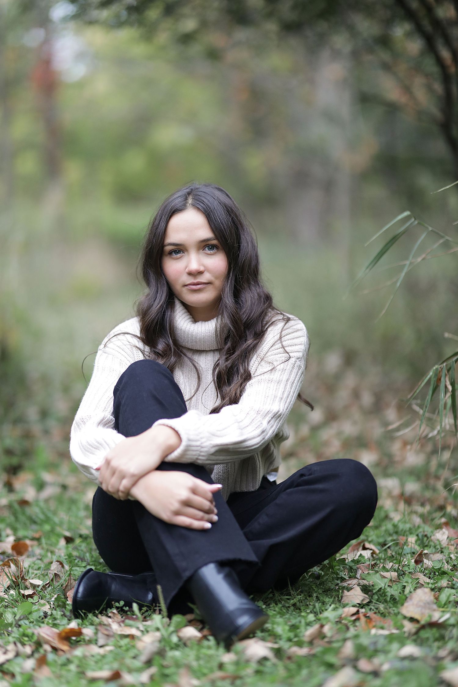 A person in a cream cable knit sweater and black pants sits cross-legged on autumn leaves in a forest setting.