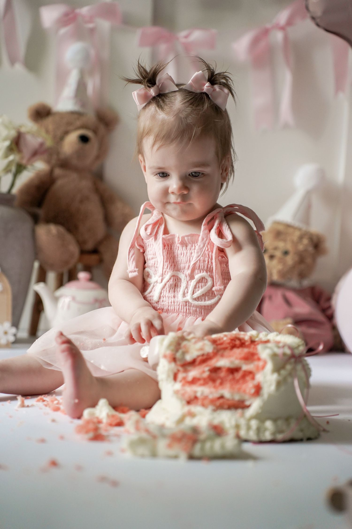 Pink cake smash birthday photo setup with teddy bears and fabric decorations against white background.
